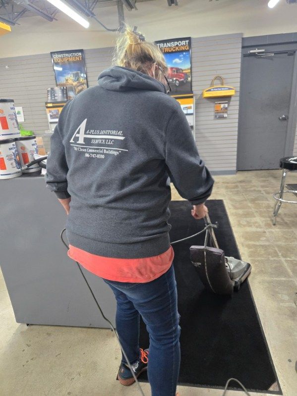 Person vacuuming carpet in a store. Gray hoodie, jeans, red shirt, and black and gray vacuum.