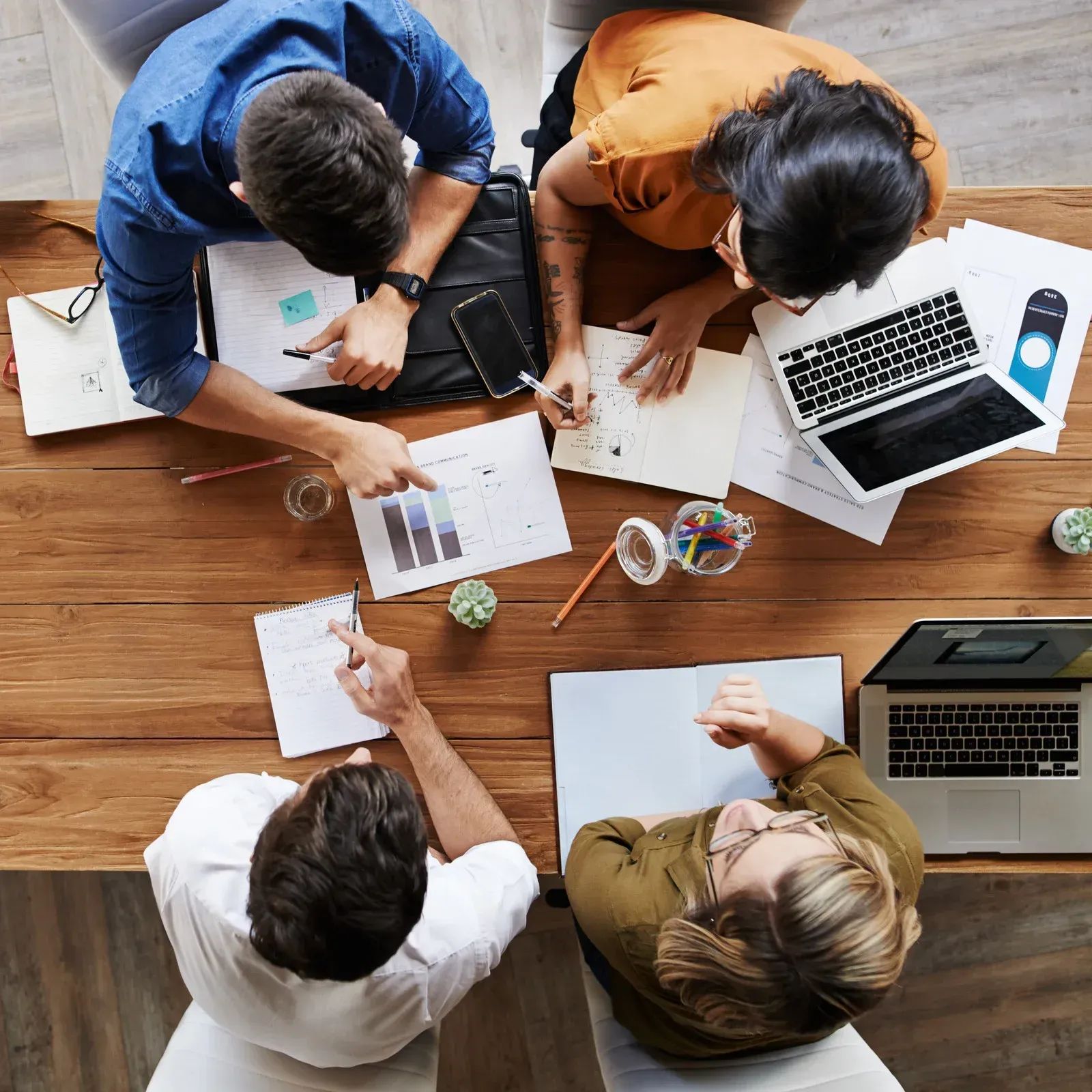Four people collaborating around a wooden table with papers, laptops, and a plant.