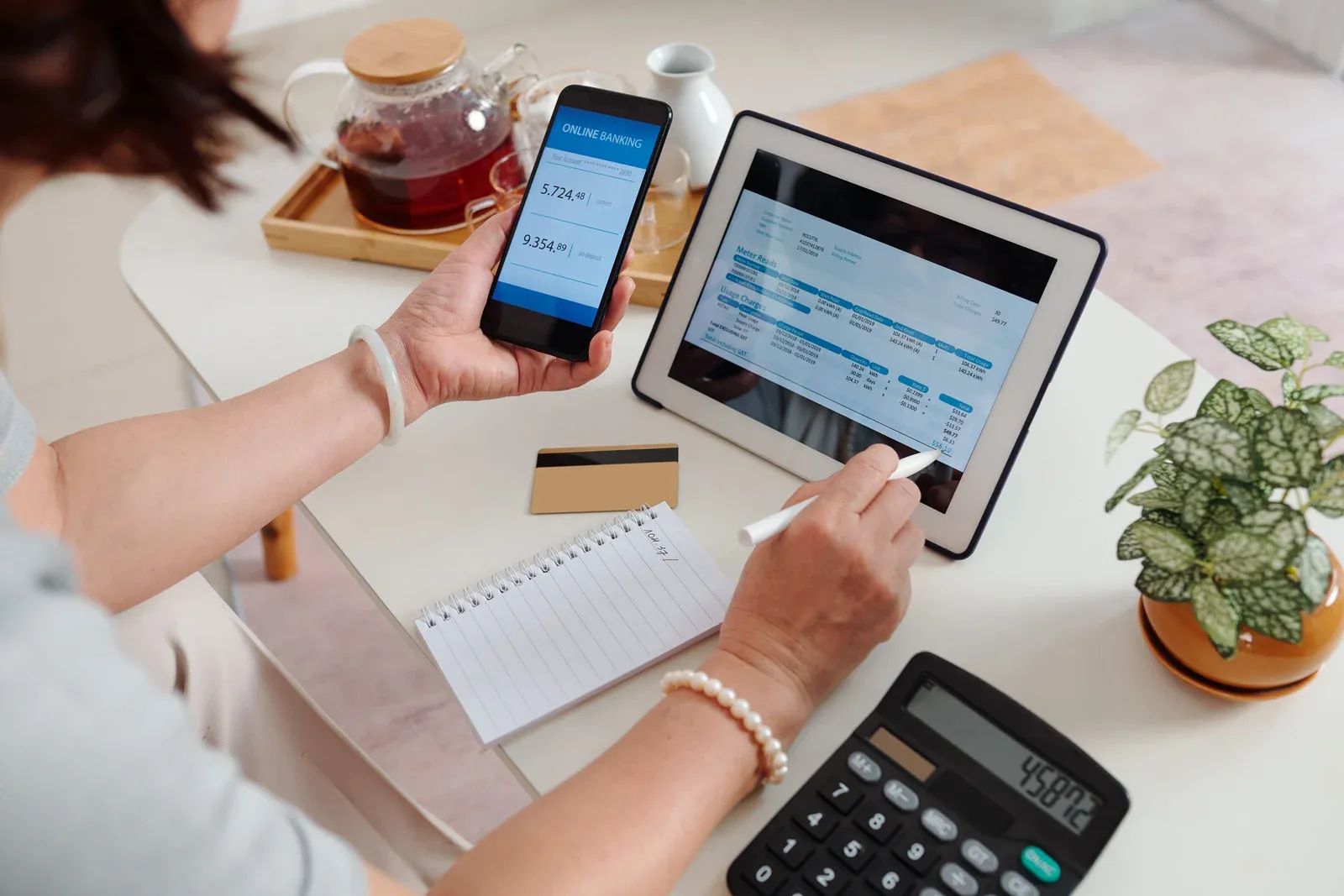 A woman is sitting at a table using a tablet and a cell phone.