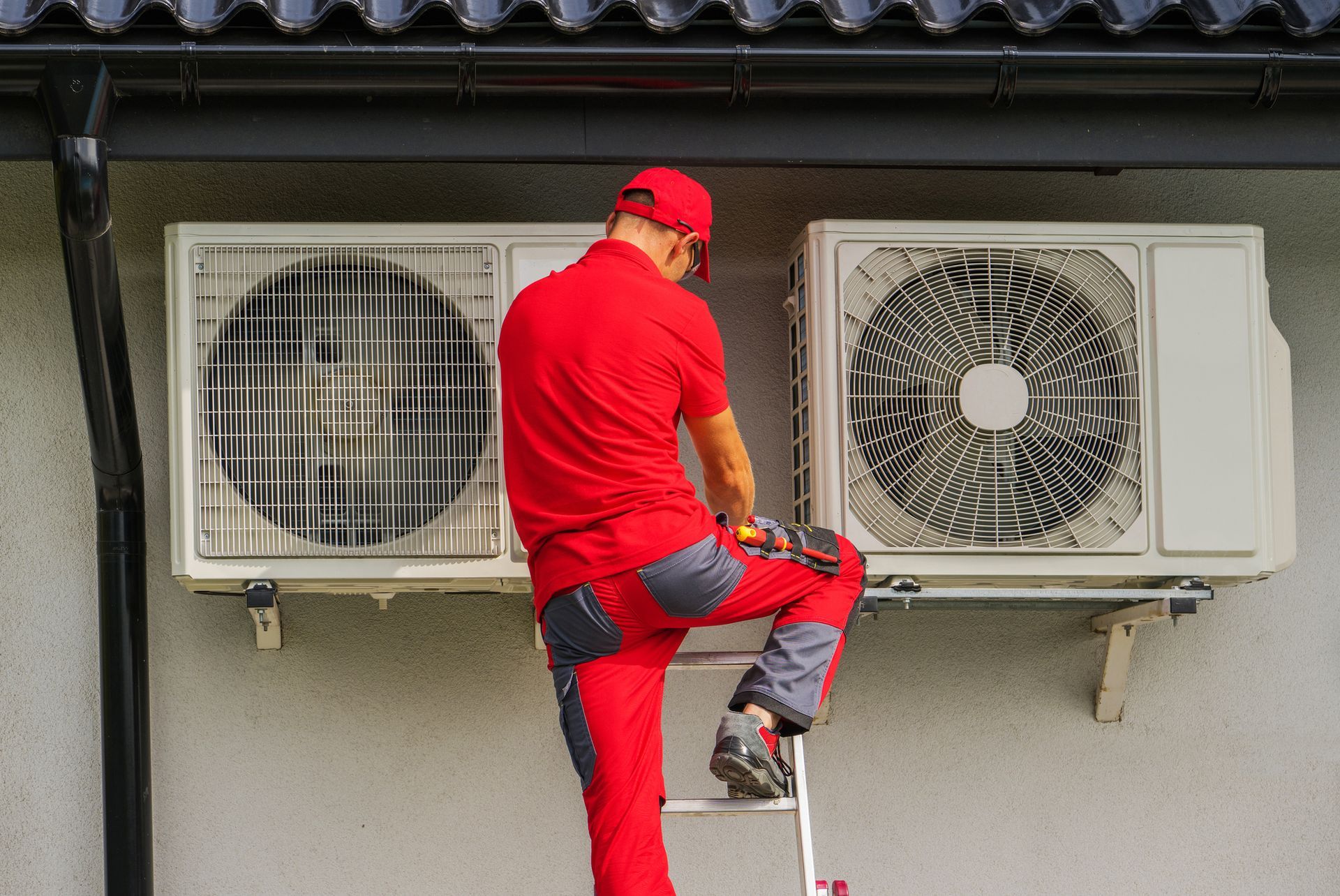 Male technician in red uniform performs residential heating repair on exterior HVAC unit.