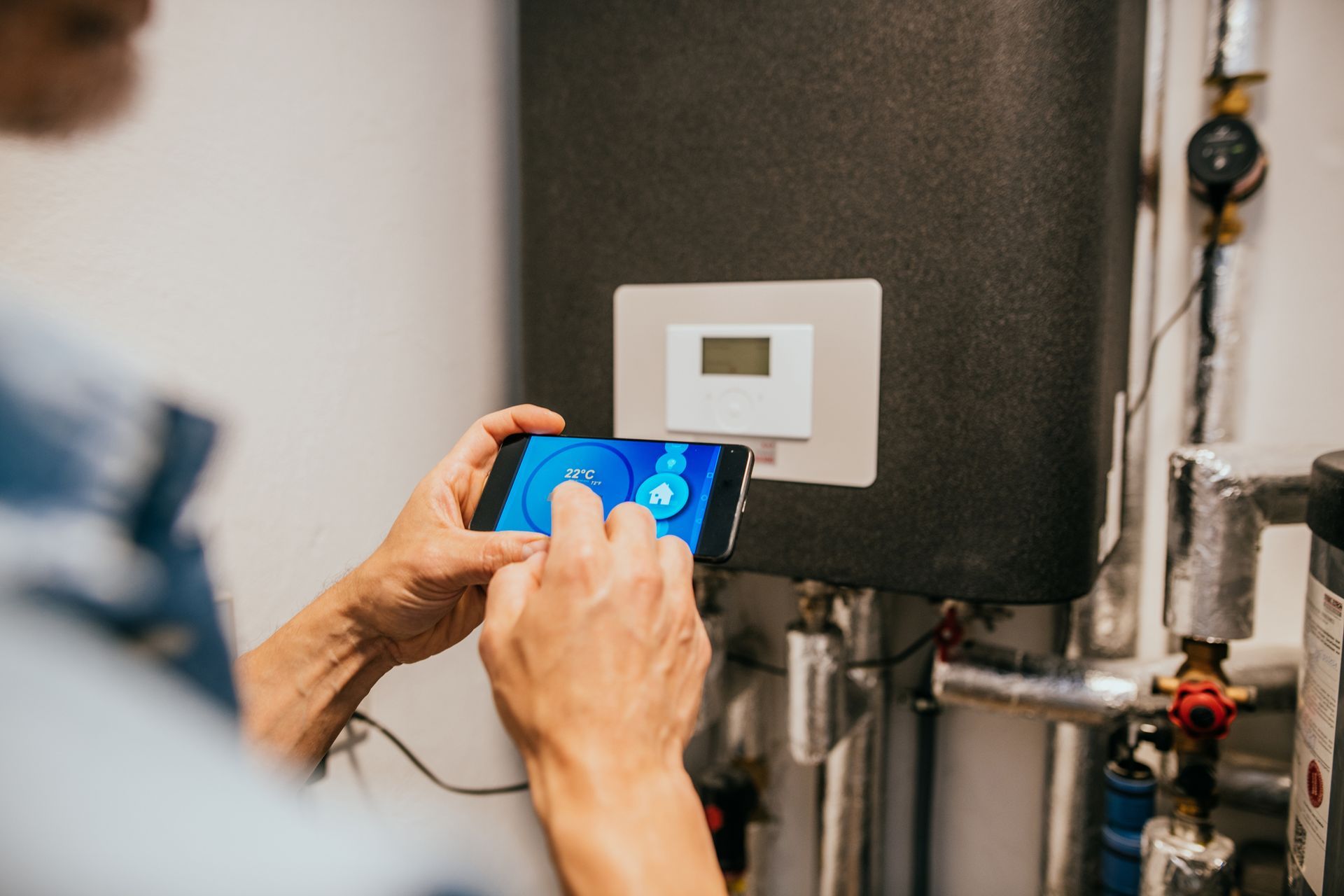 Person adjusting digital thermostat using smartphone near heating system