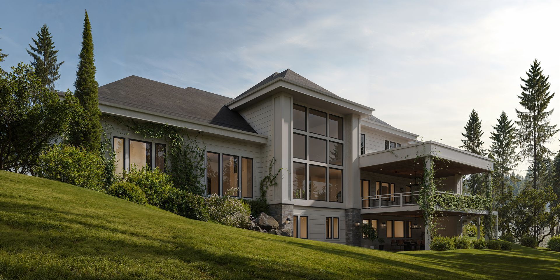 A large, modern house with tall windows and a stone patio sits on a green, sloped lawn surrounded by tall pine trees.