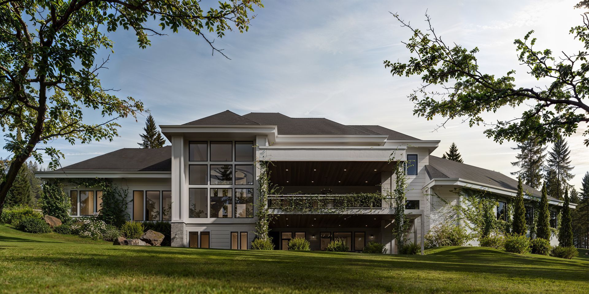 A modern, light-colored house with a large windowed wing and a covered terrace, situated on a lush, sunny green lawn.