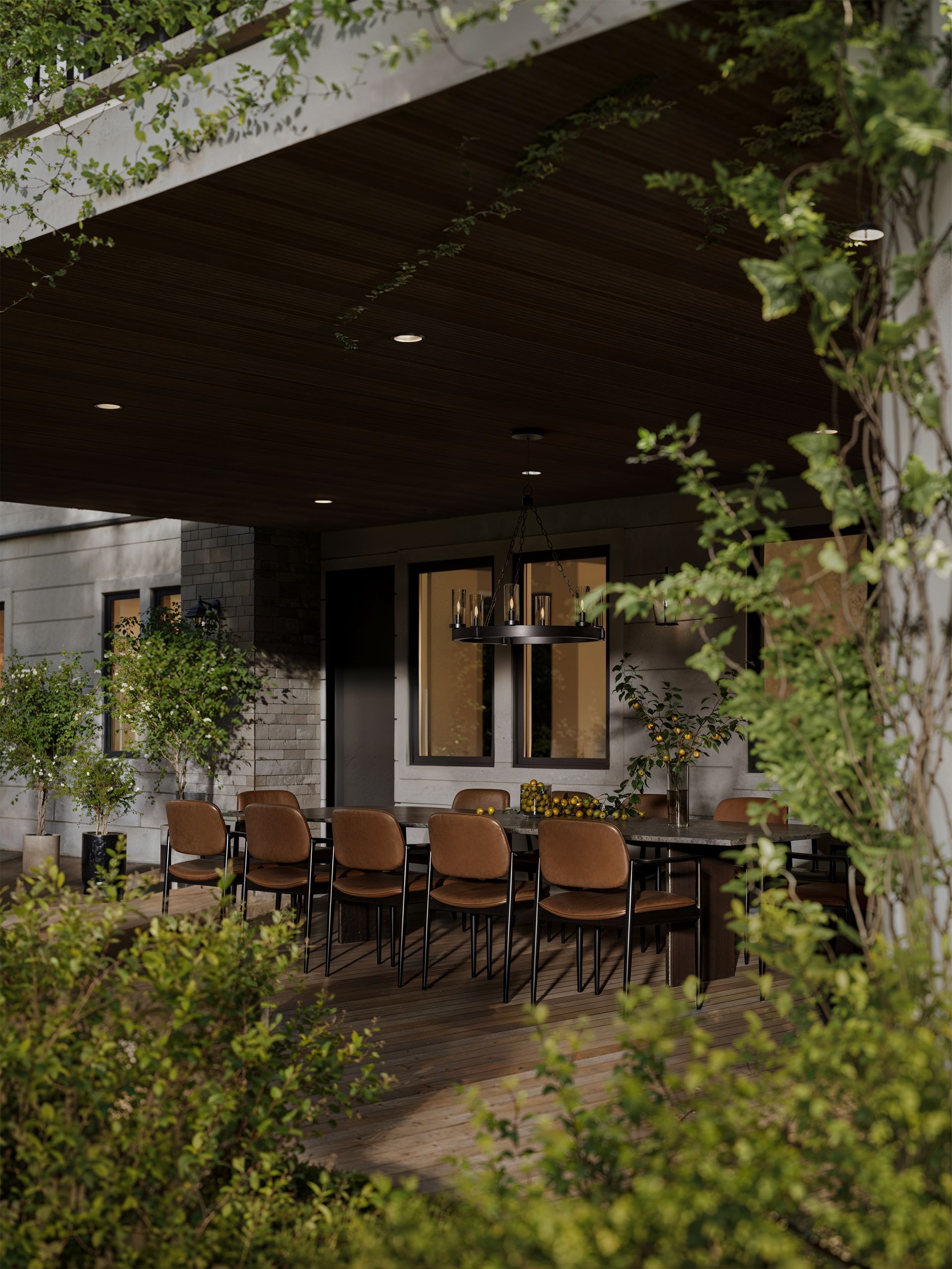 A covered outdoor patio with a long dining table, brown chairs, and warm lighting, surrounded by greenery.
