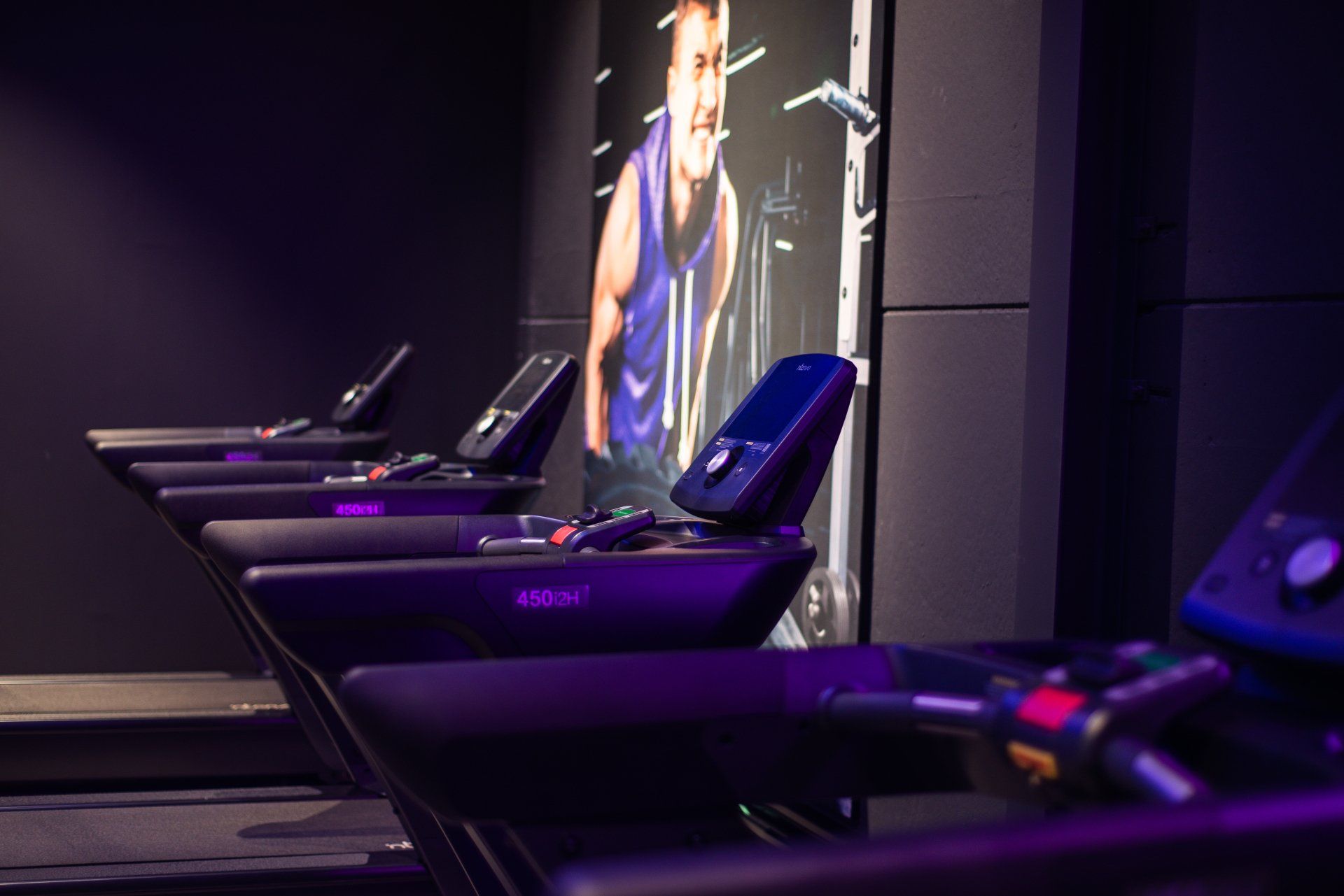 Row of treadmills in a dim gym, illuminated in purple light, with a poster of a singer in the background.