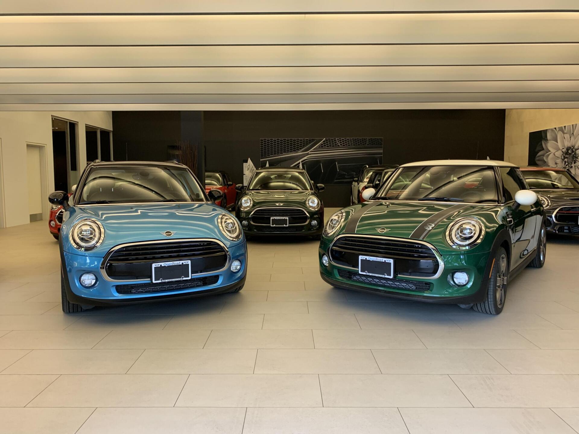 Several Mini Coopers in a showroom: blue, green, and black cars are displayed, with a black wall backdrop.