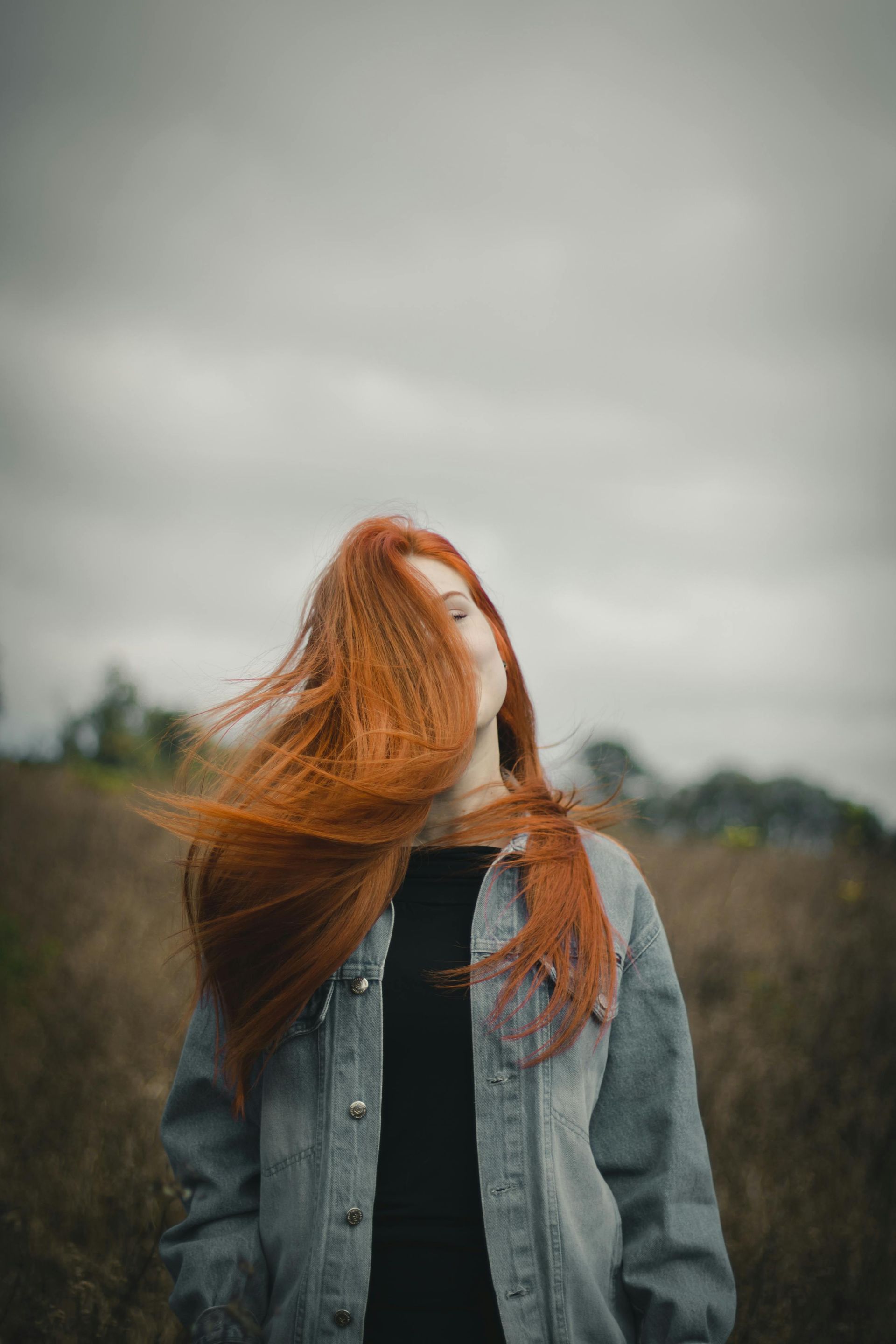 A woman with long red hair is standing in a field with her hair blowing in the wind.