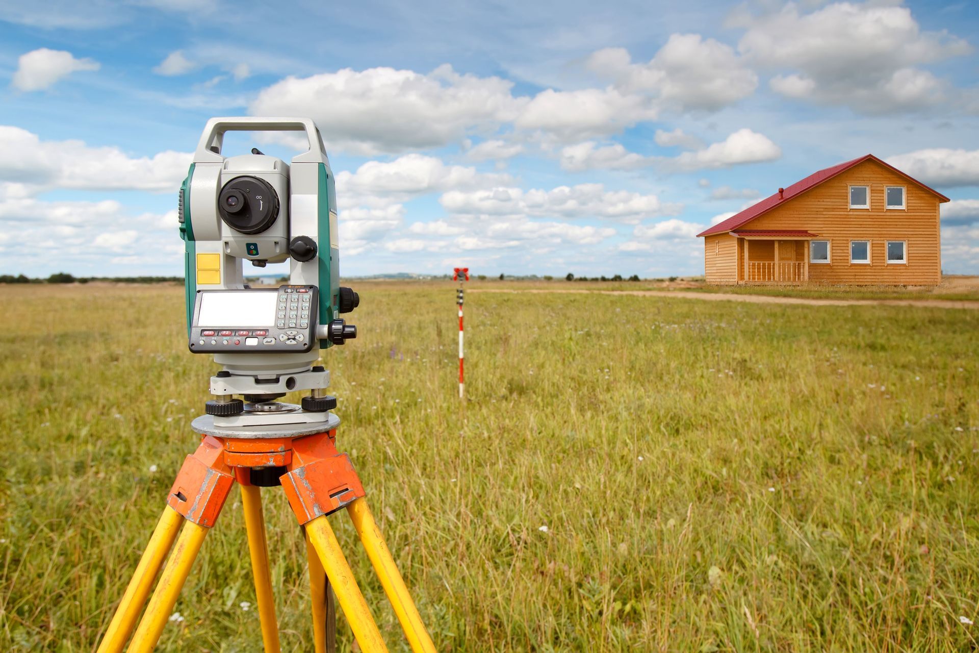 Surveying equipment on a tripod in a field, a house in the background, a blue and cloudy sky.