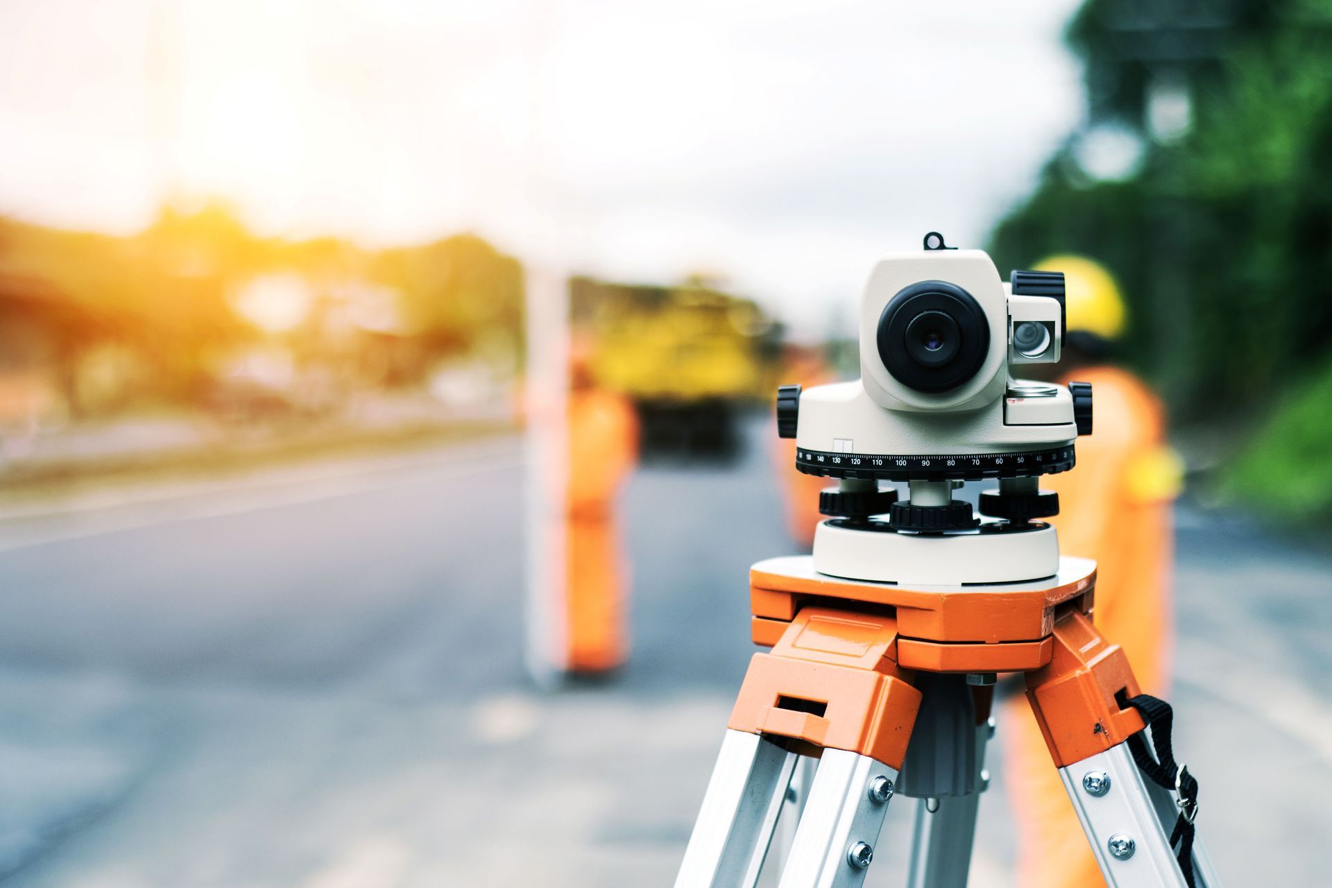 Surveying instrument on a tripod, orange and white, roadside setting, with workers in orange vests blurred in the background.