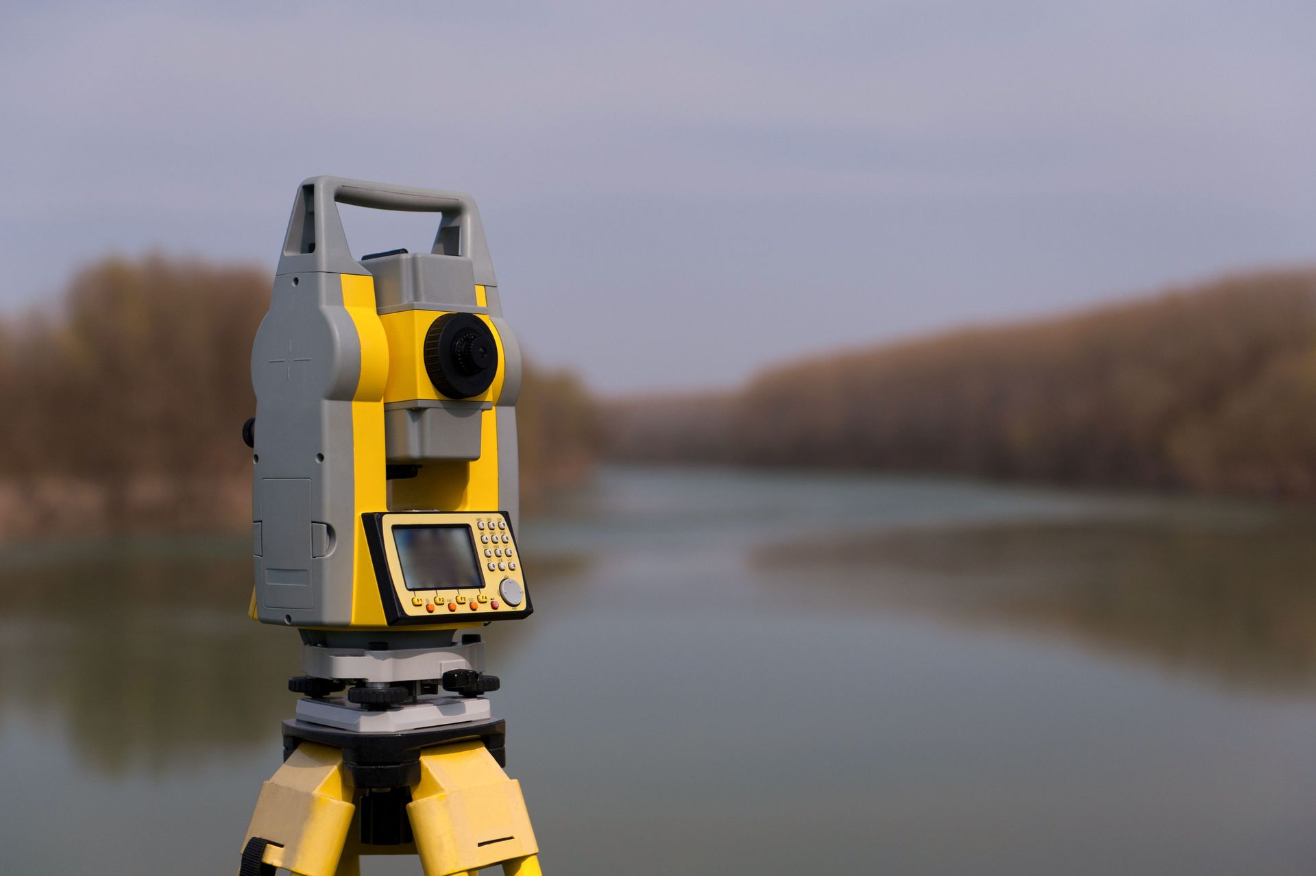 Yellow and gray surveying instrument on a tripod, overlooking a river with trees in the background.