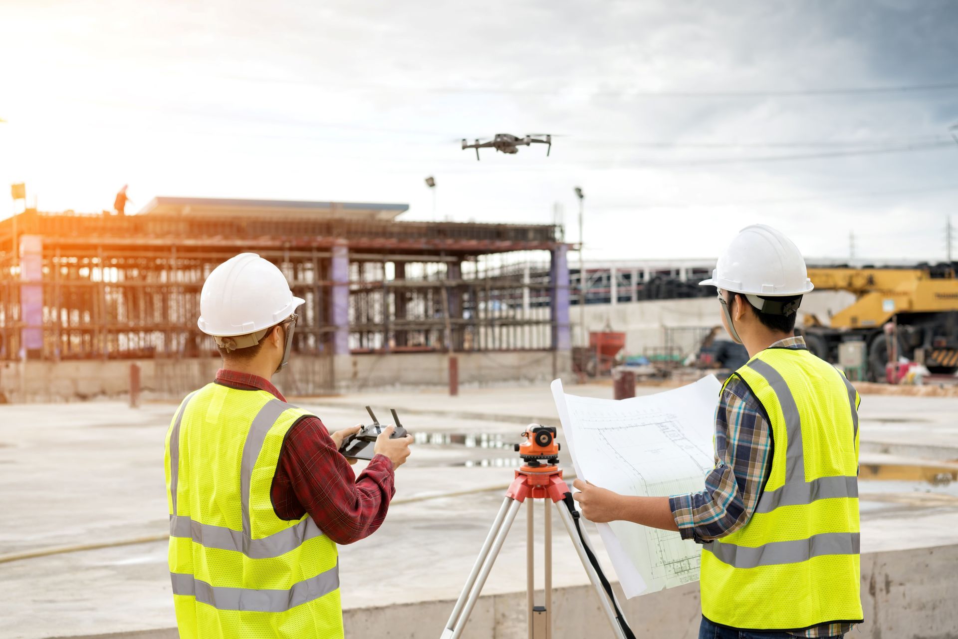 Two construction workers in safety vests and hard hats operating a drone at a construction site.