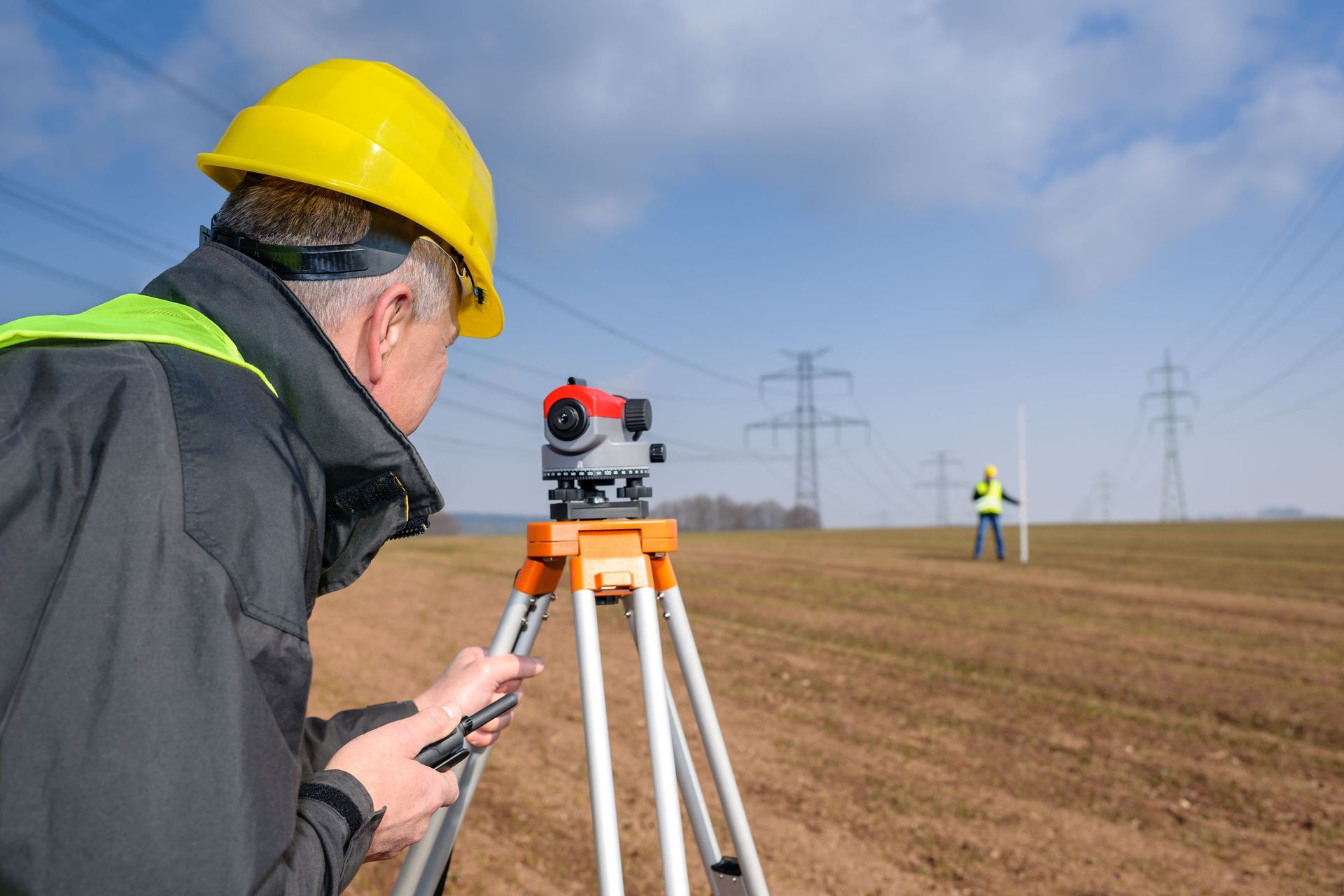 Surveyor using a level, sighting a worker holding a measurement rod in a field near power lines.