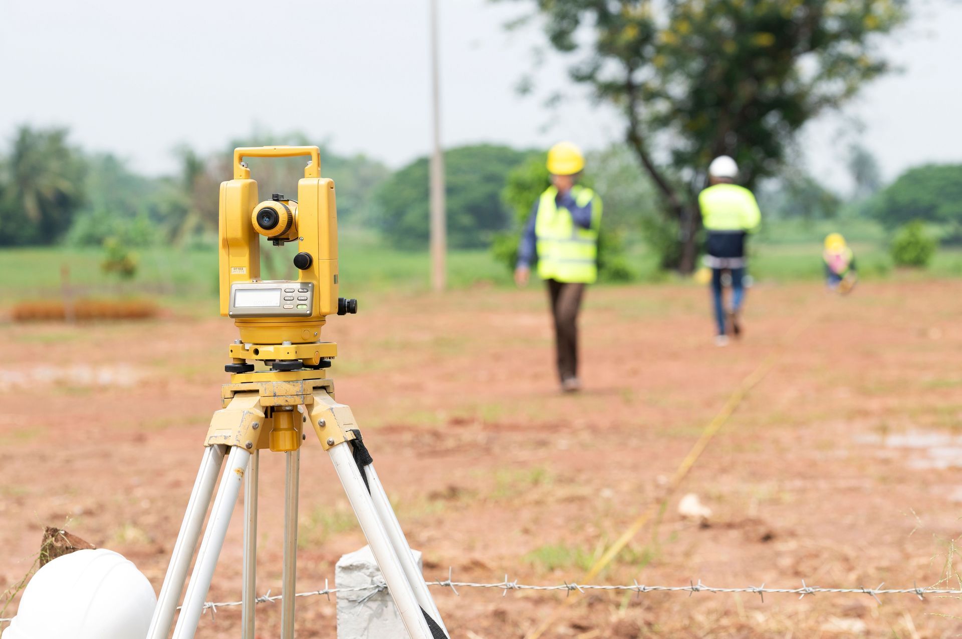 Yellow theodolite on tripod at a construction site; two workers in vests in the background.