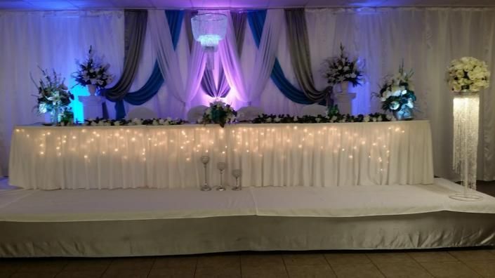 Decorated wedding stage with white drapes, purple lighting, floral arrangements, and twinkling string lights
