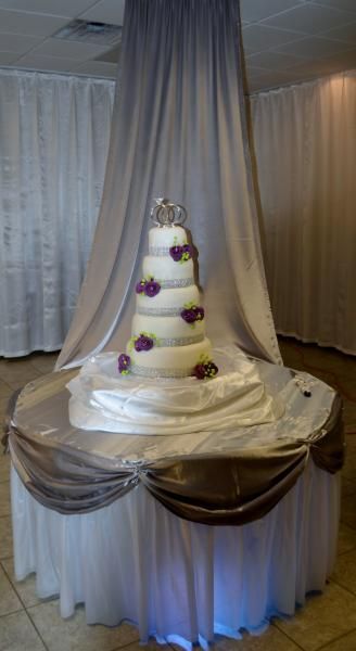 White tiered wedding cake with purple flowers on a draped round table under a sheer canopy indoors