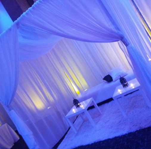 Blue-lit canopy lounge with sheer drapes, white bench, small tables, and candles on a fuzzy rug