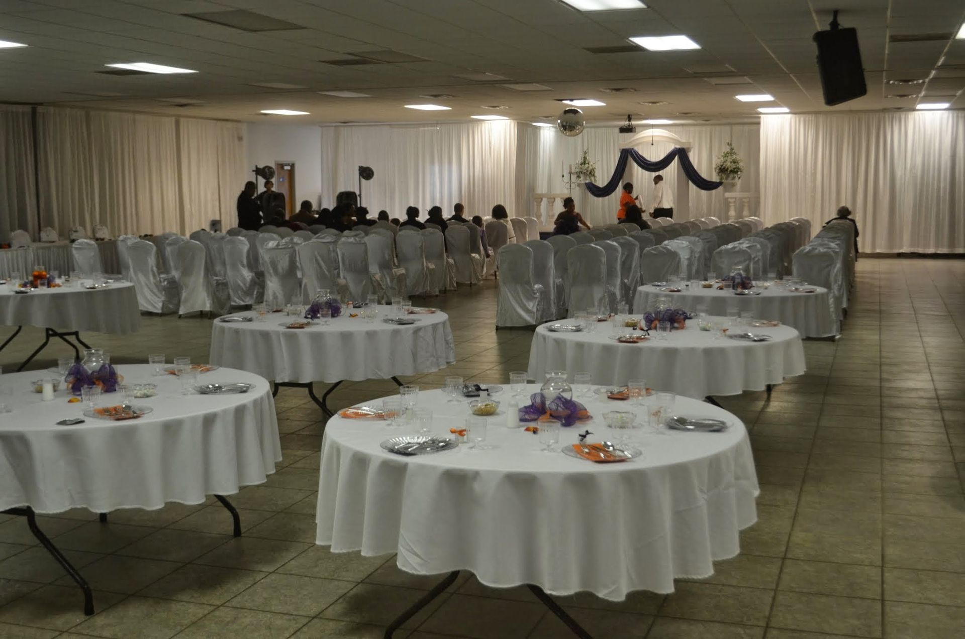 Banquet hall with round, white-draped tables set for an event and guests gathered at the back stage.