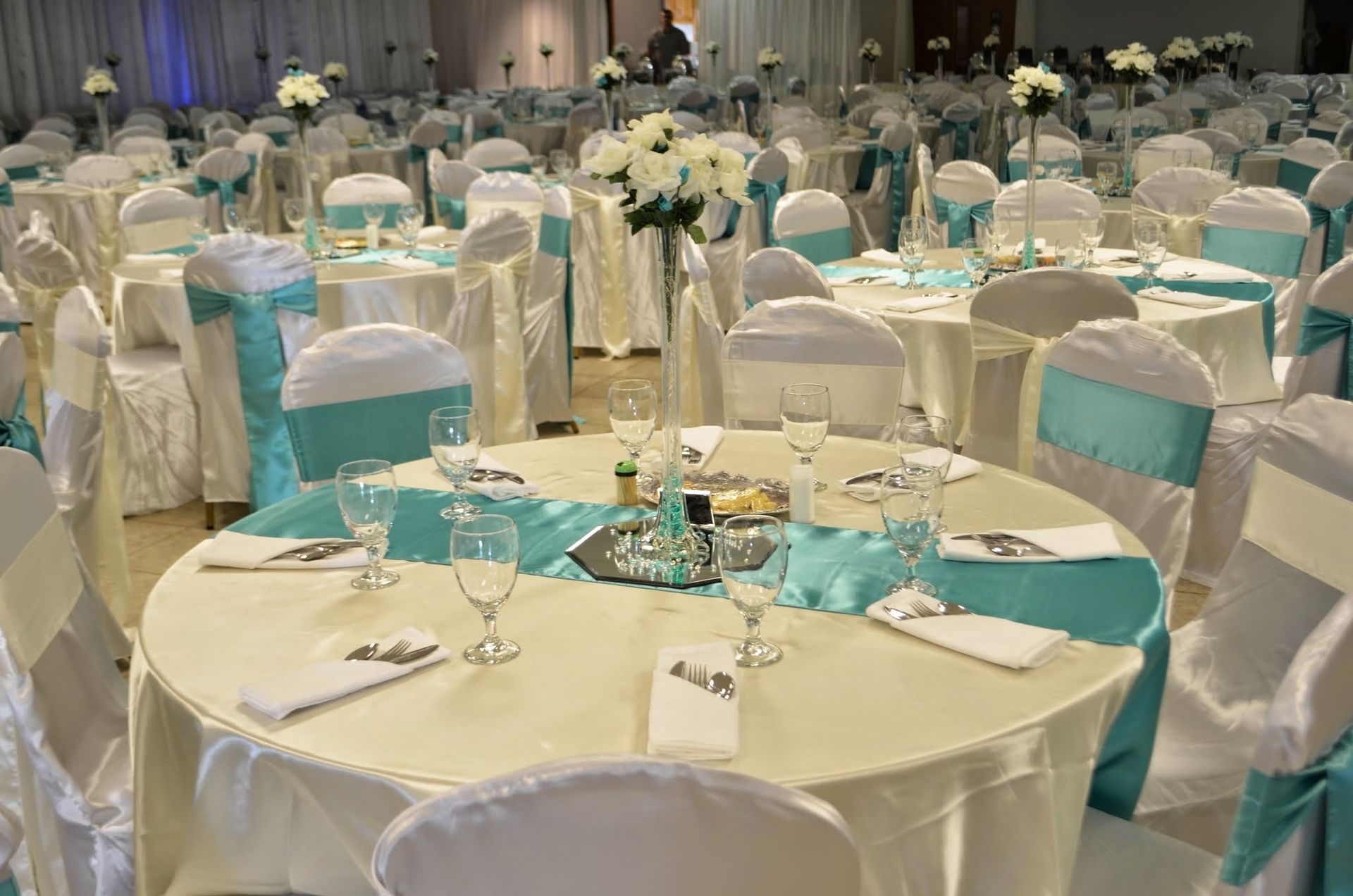 Round banquet tables set for an event, with white chair covers and turquoise table runners in a decorated hall