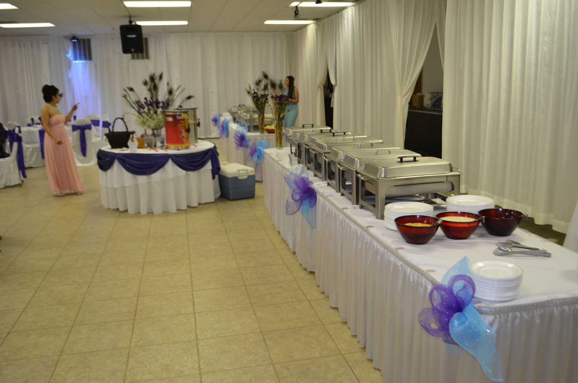 Decorated banquet hall with buffet tables, white drapes, blue bows, and a woman in a pink dress near the back