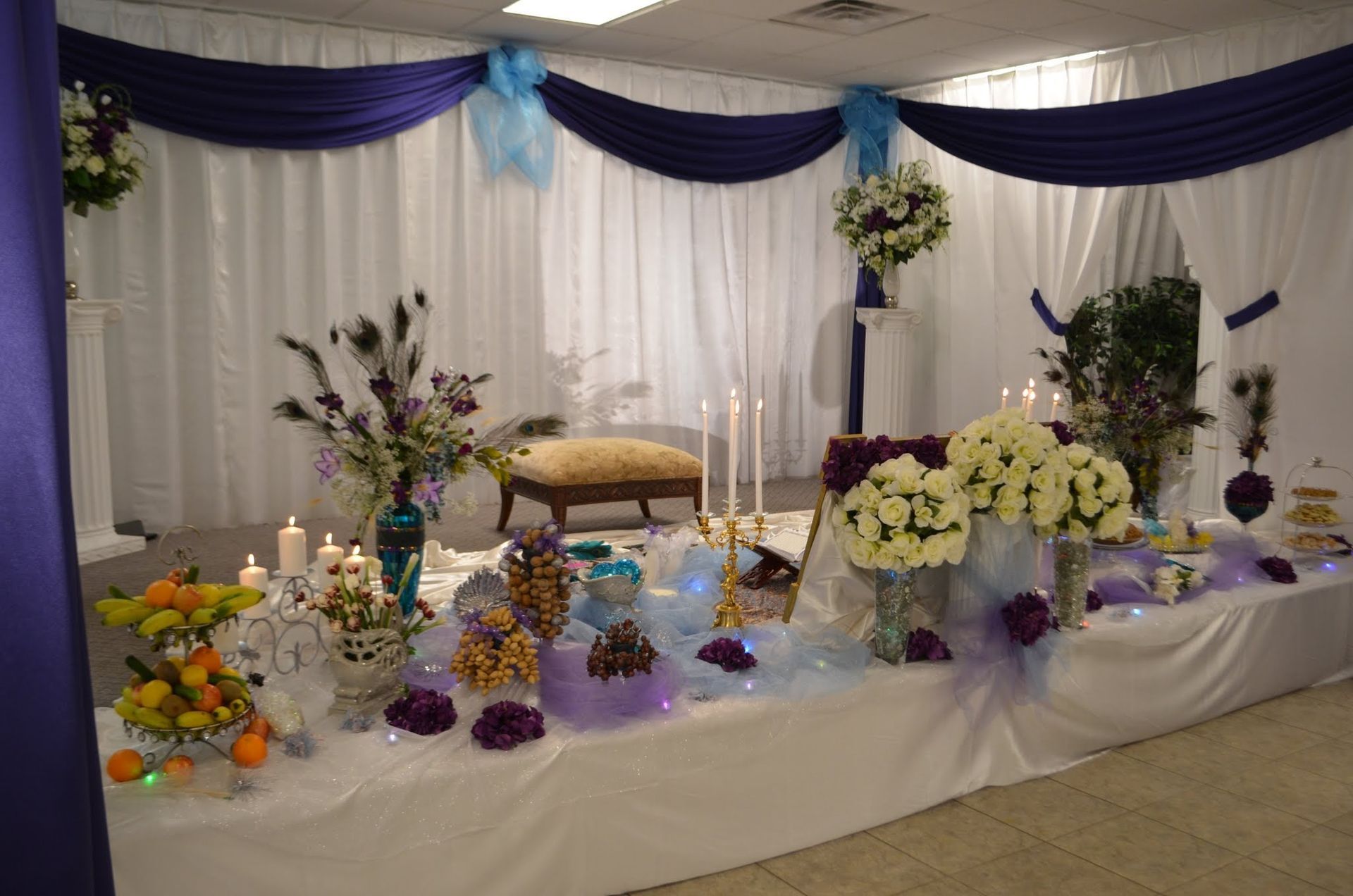 Decorated banquet table with white and purple drapes, floral arrangements, candles, and fruit displays