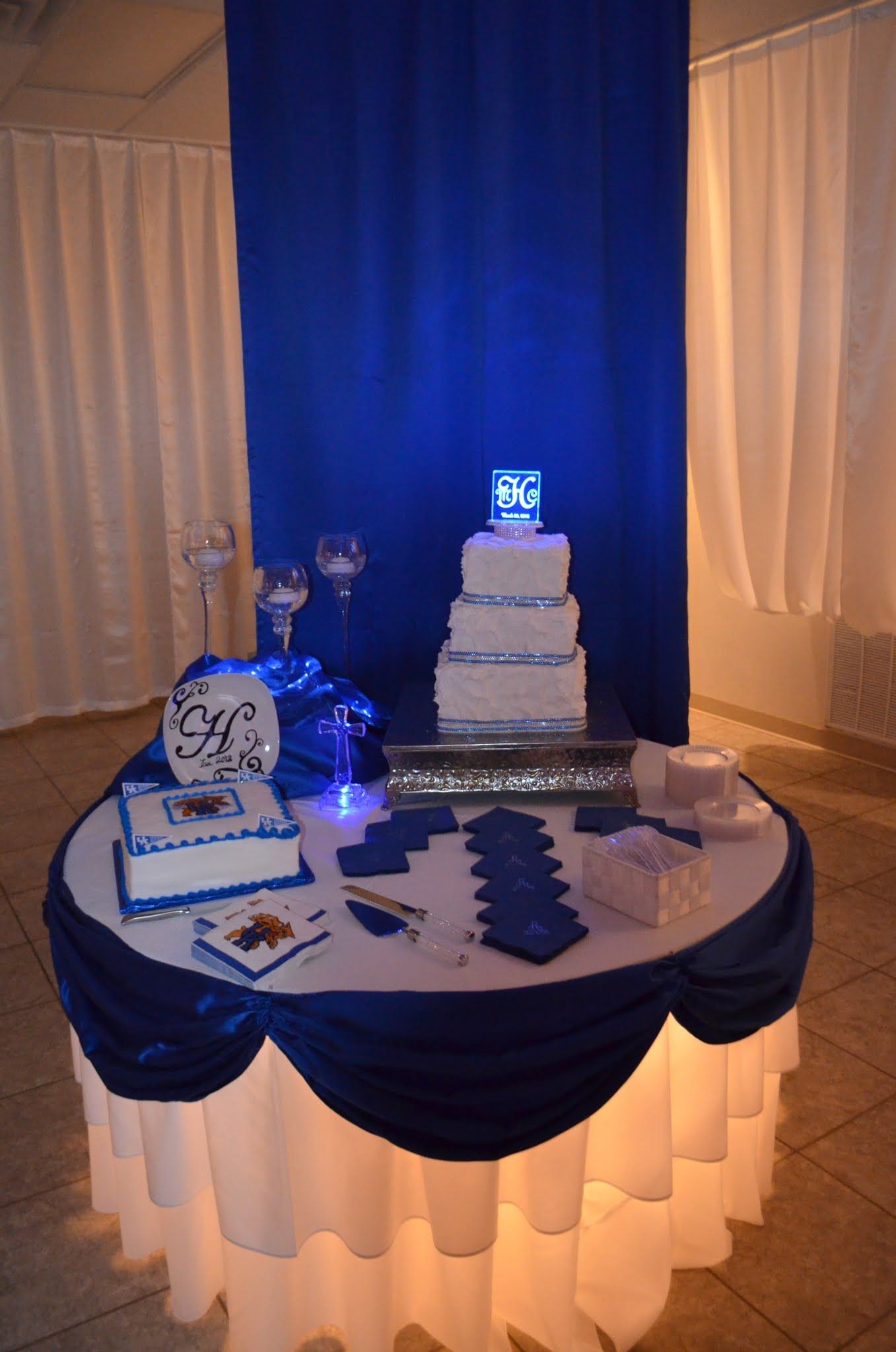 Decorated dessert table with a tiered white cake, blue drapes, and glowing blue accents at an event