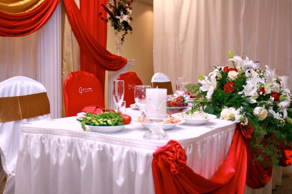 Banquet table with white cloth, red drapes, floral centerpiece, and place settings in a decorated hall