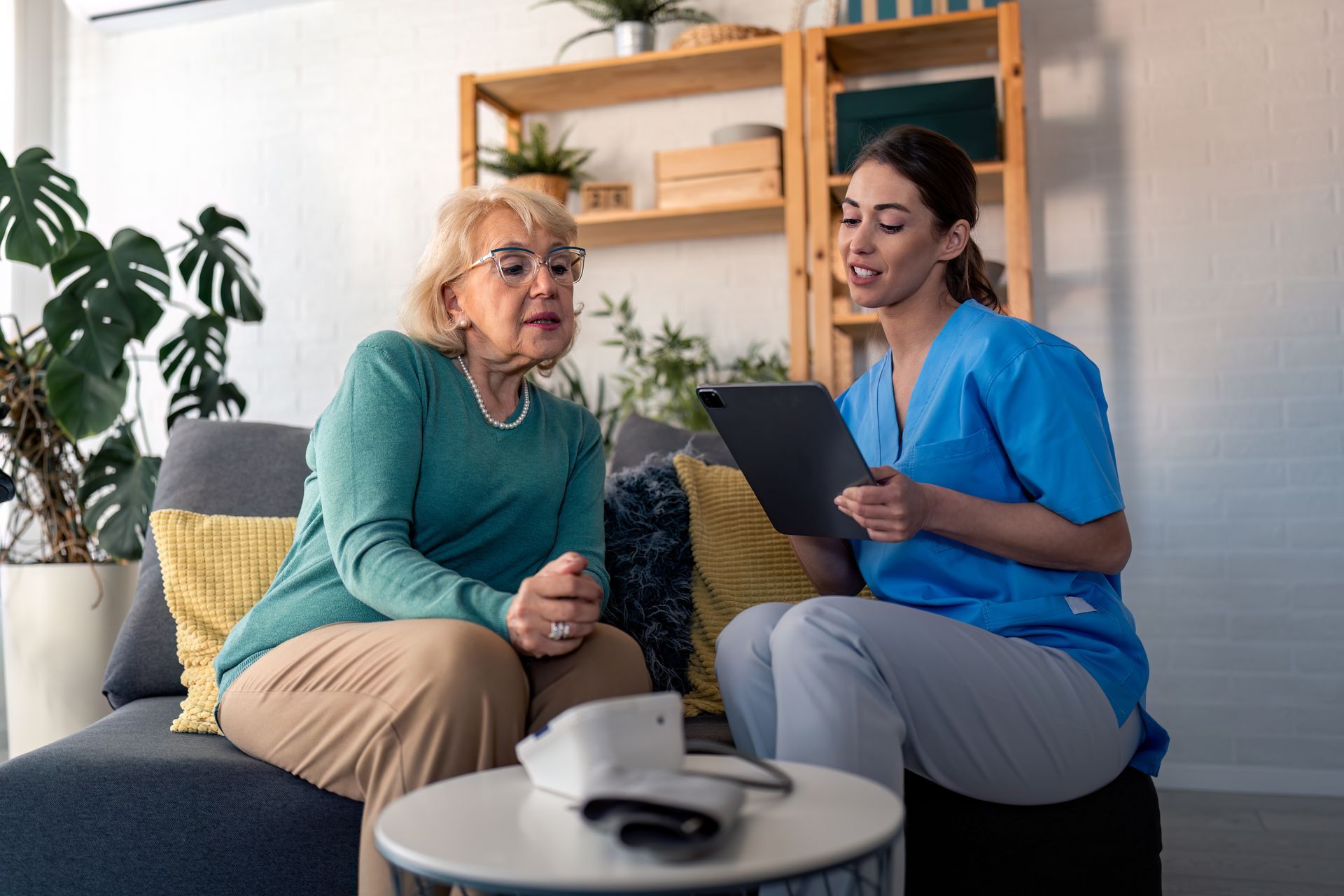 Nurse reviewing a tablet with a seated patient in a bright living room, with a blood pressure monitor on the table Nurse reviewing a tablet with a seated patient in a bright living room, with a blood pressure monitor on the table
