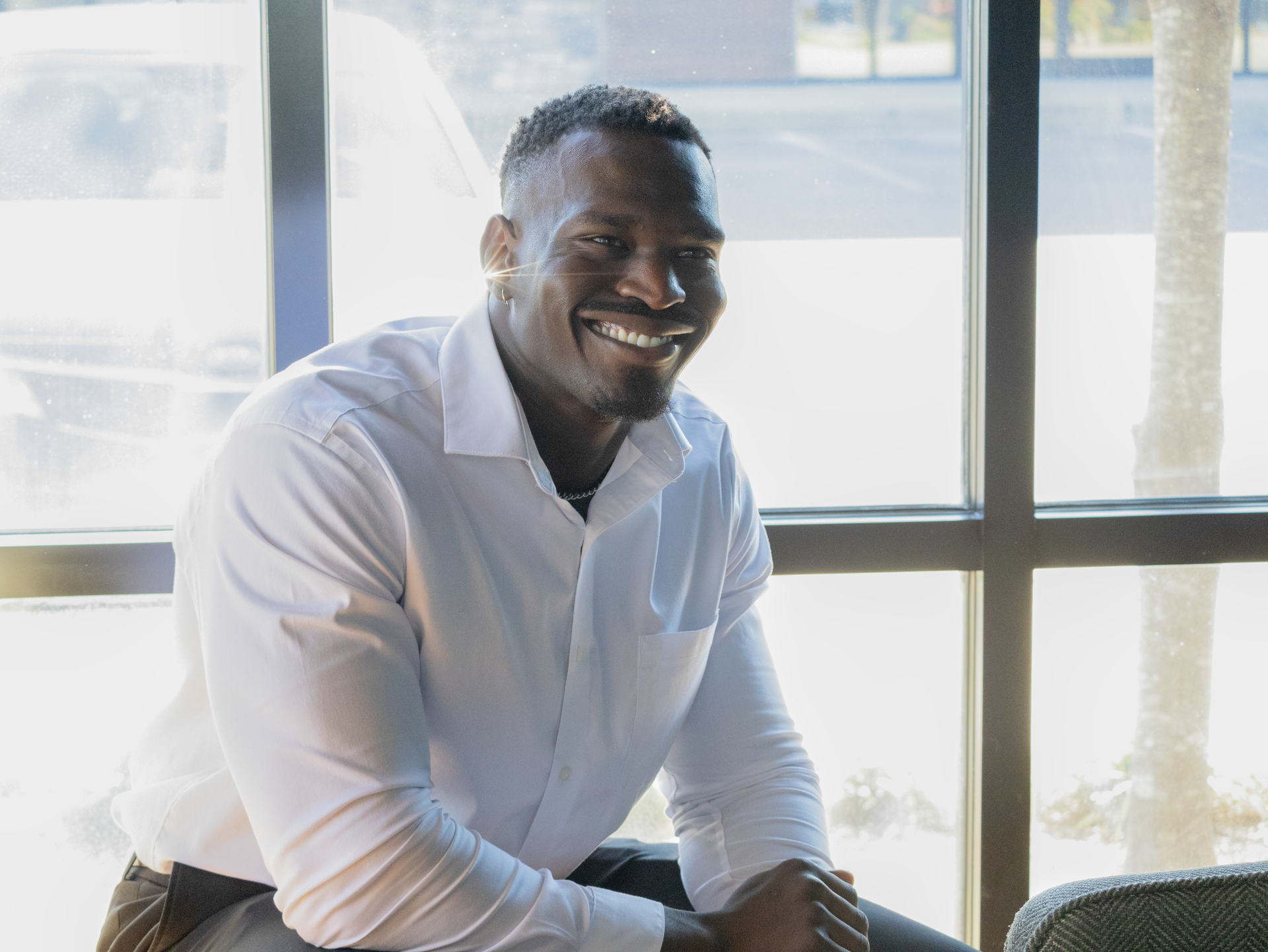 A man in a white shirt is sitting in front of a window and smiling.