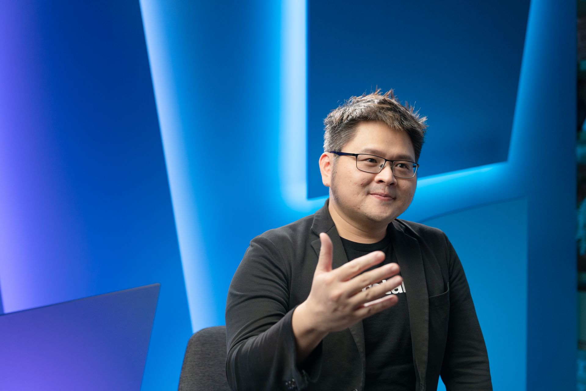 A man in a suit and glasses is sitting in front of a blue wall.