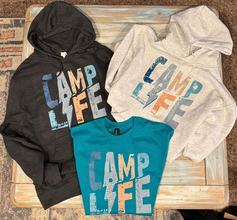 Three camp life sweatshirts are sitting on a wooden table