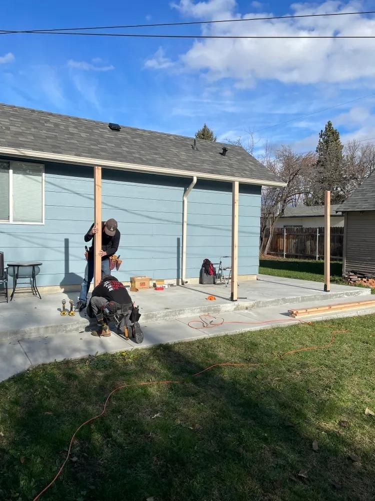 Person working on a patio beside a blue house, with a dog nearby and a grassy yard in front.