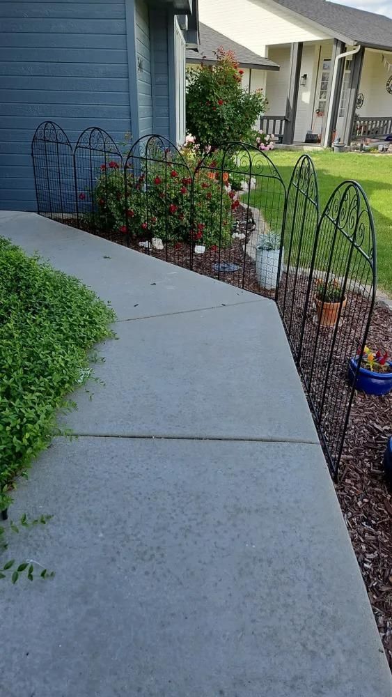 Curved concrete walkway beside a blue house, bordered by flower beds and small garden arches.