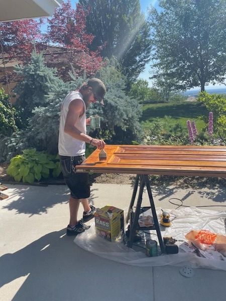 Person staining a wooden table outdoors in a sunny garden, with tools and supplies nearby