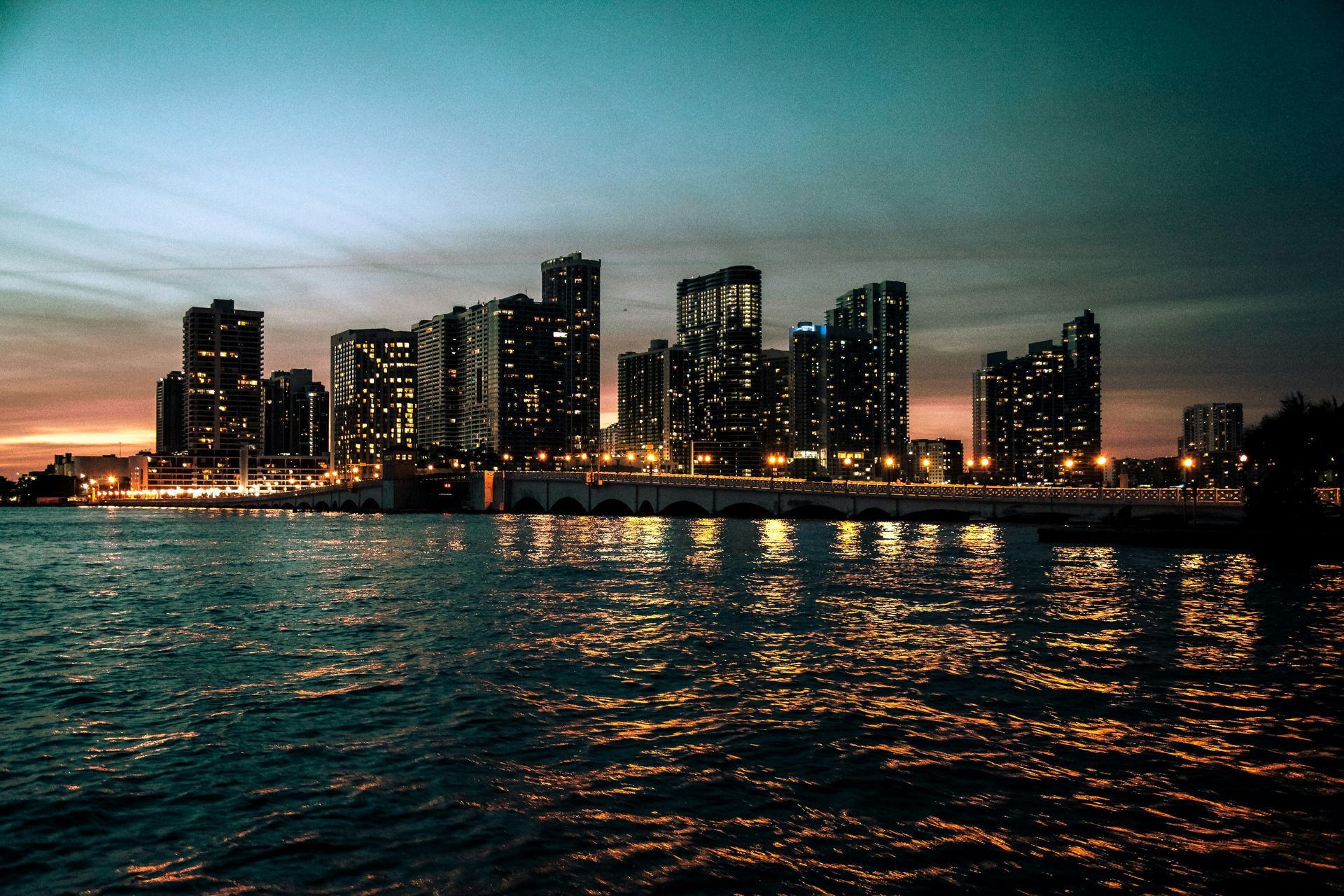 City skyline at dusk reflected on rippling water, with glowing buildings and a colorful sunset sky