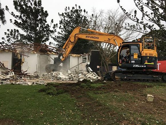 A yellow excavator is demolishing a building in a field.
