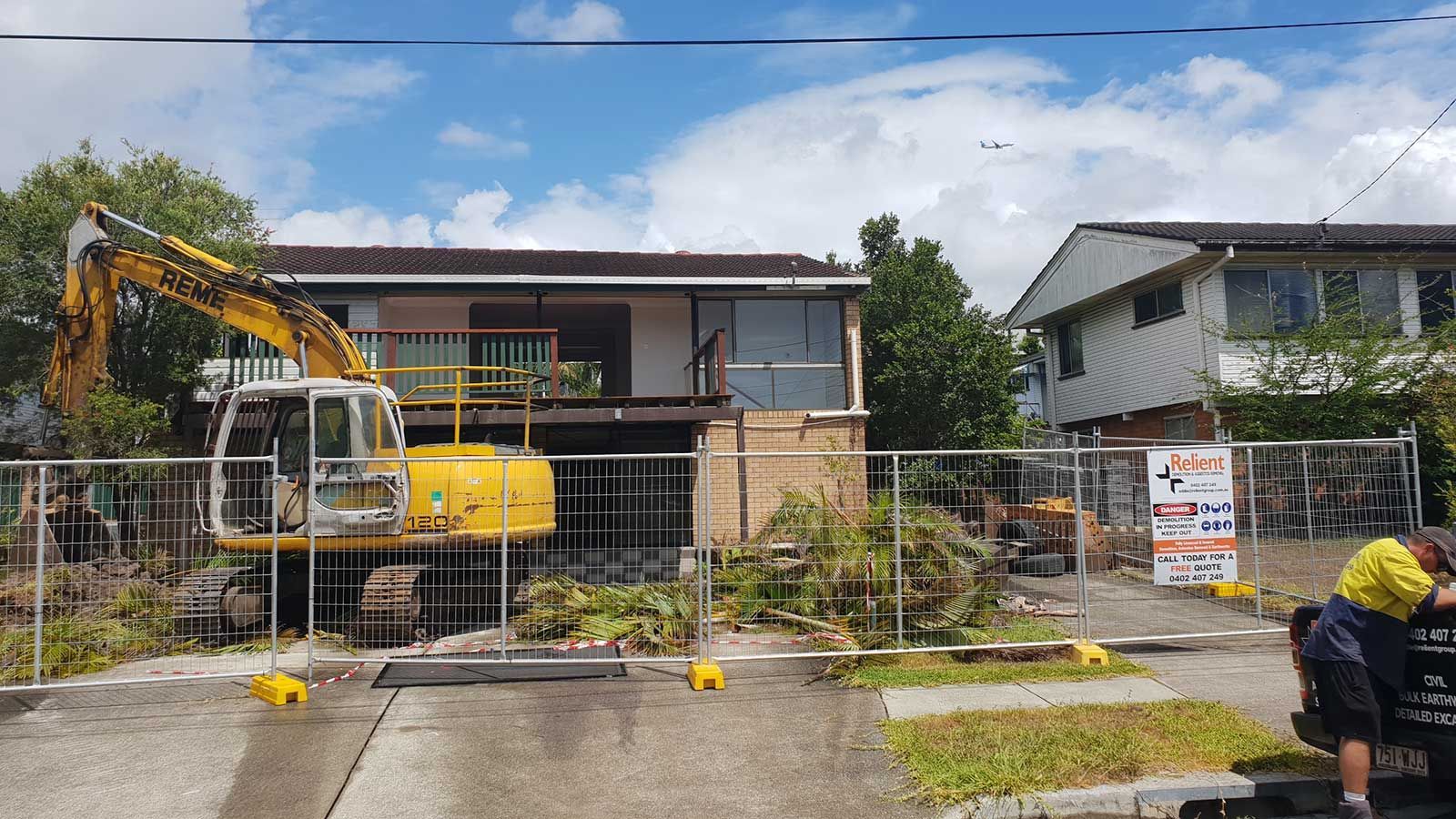 A yellow excavator is demolishing a house behind a fence.