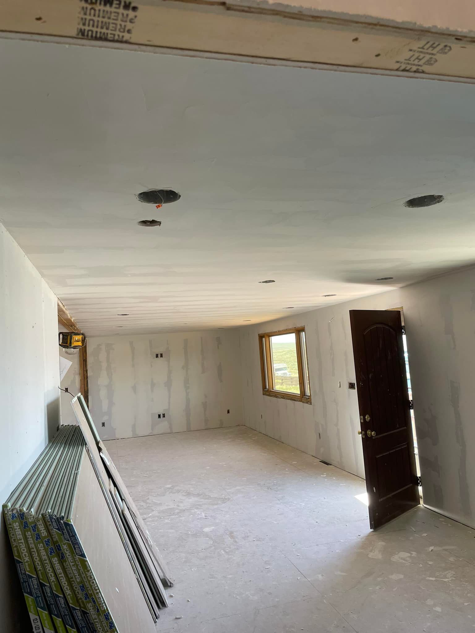 Room under construction, drywall on walls and ceiling. Sheetrock stacked. Window and door visible.