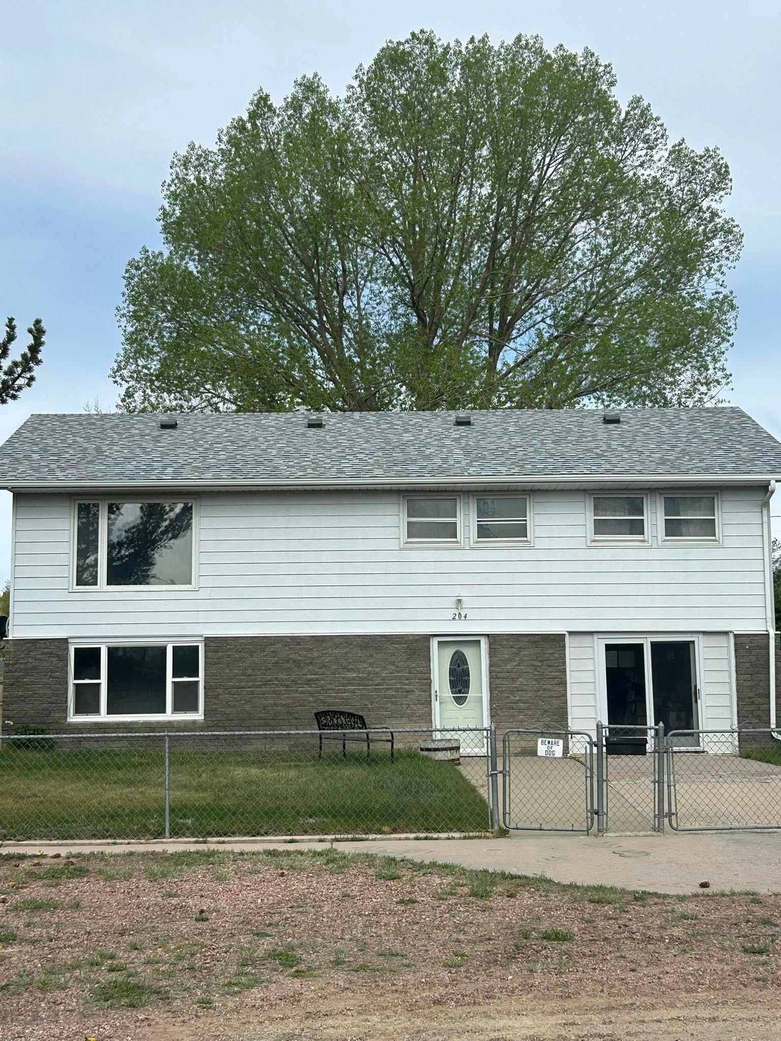 Two-story white house with brick facade, large tree behind, gray roof, chain link fence.