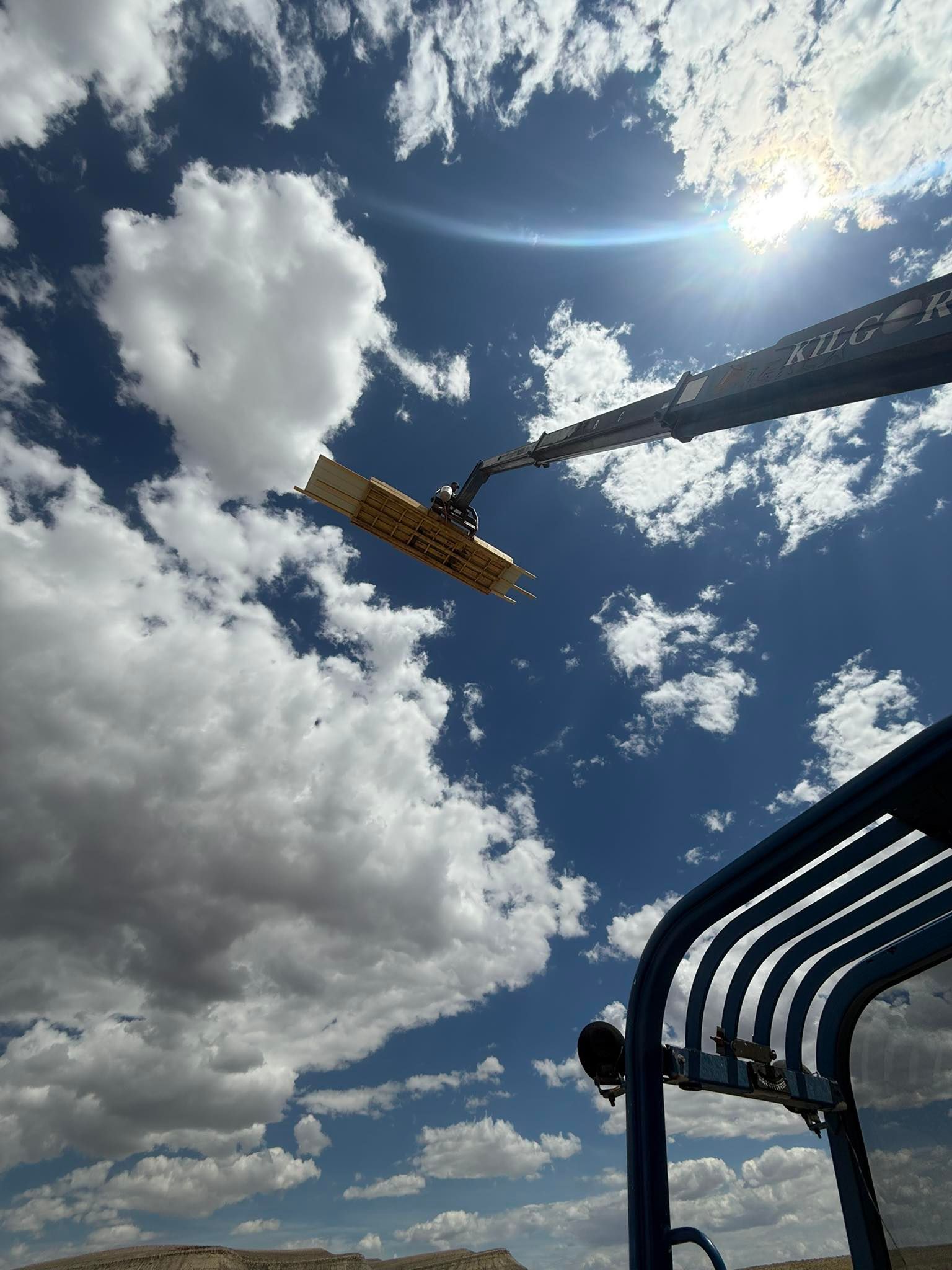 Sky view with a boom lift reaching for the sky; blue sky, white clouds, and bright sun.