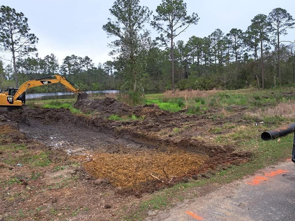 A yellow excavator digs in a muddy field next to a road, clearing brush and earth.