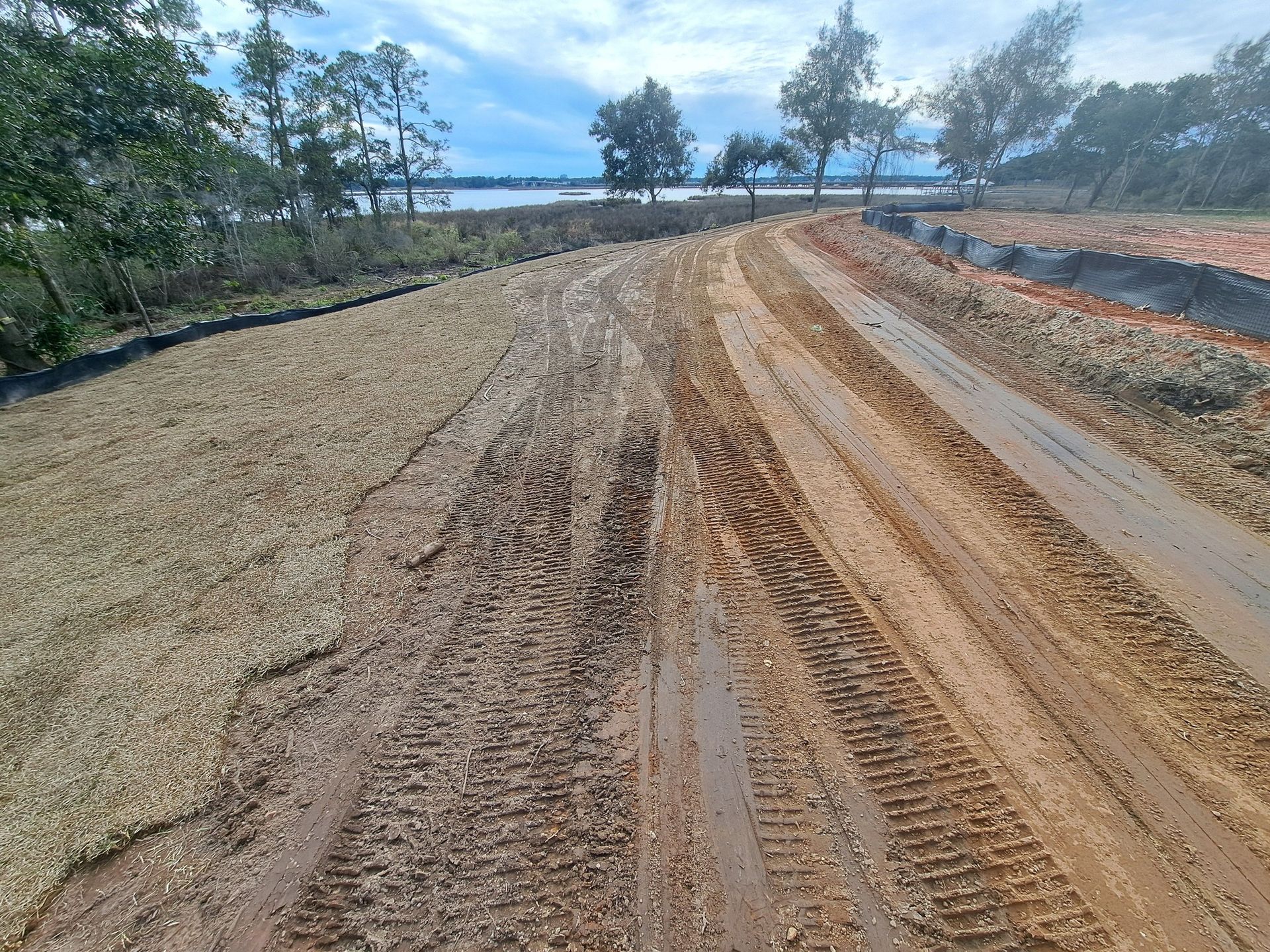 Muddy dirt road with tire tracks leading towards a lake, flanked by grassy embankments and trees .