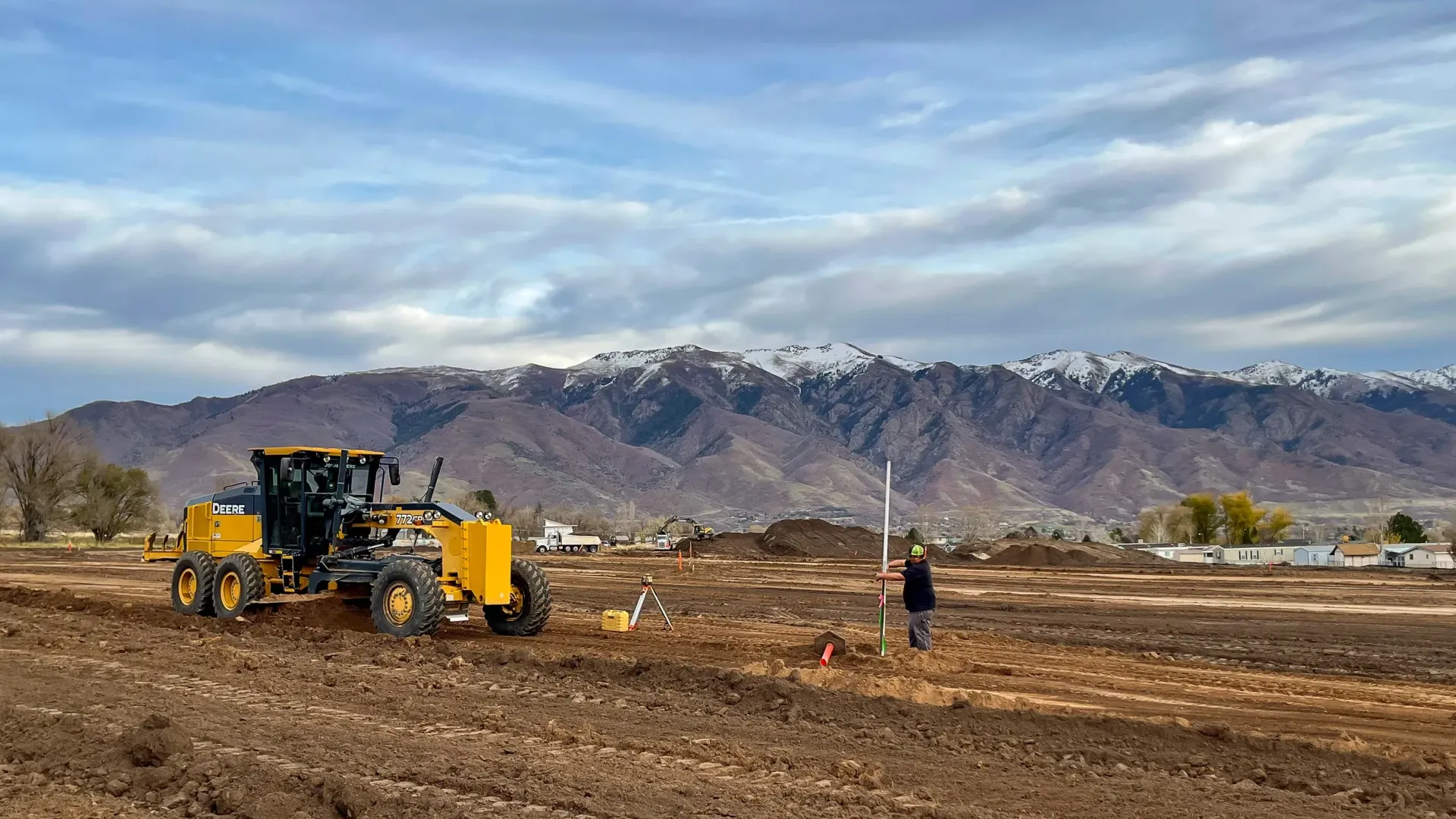 A yellow tractor is plowing a dirt field with mountains in the background.