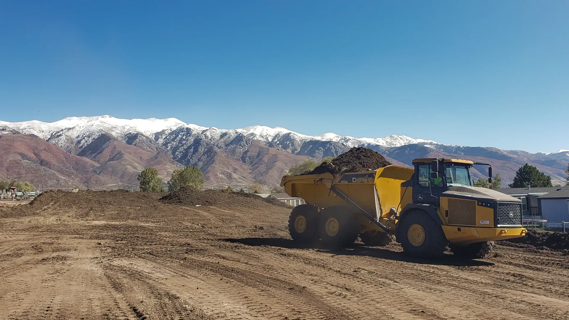 A yellow dump truck is driving through a dirt field with mountains in the background.
