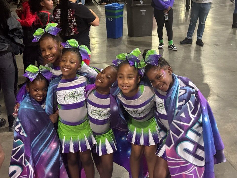 Six cheerleaders in purple-and-green uniforms pose together in a lobby, smiling with bows and capes.
