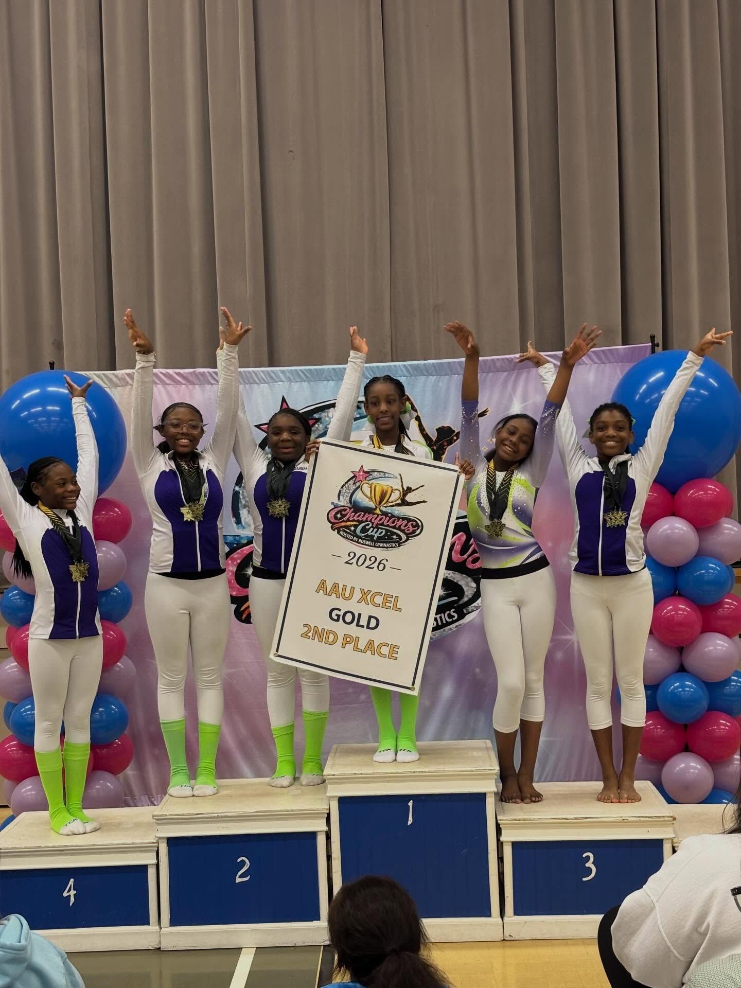 Gymnastics team on a podium holding medals and a trophy, with balloons and place numbers 1, 2, and 3.