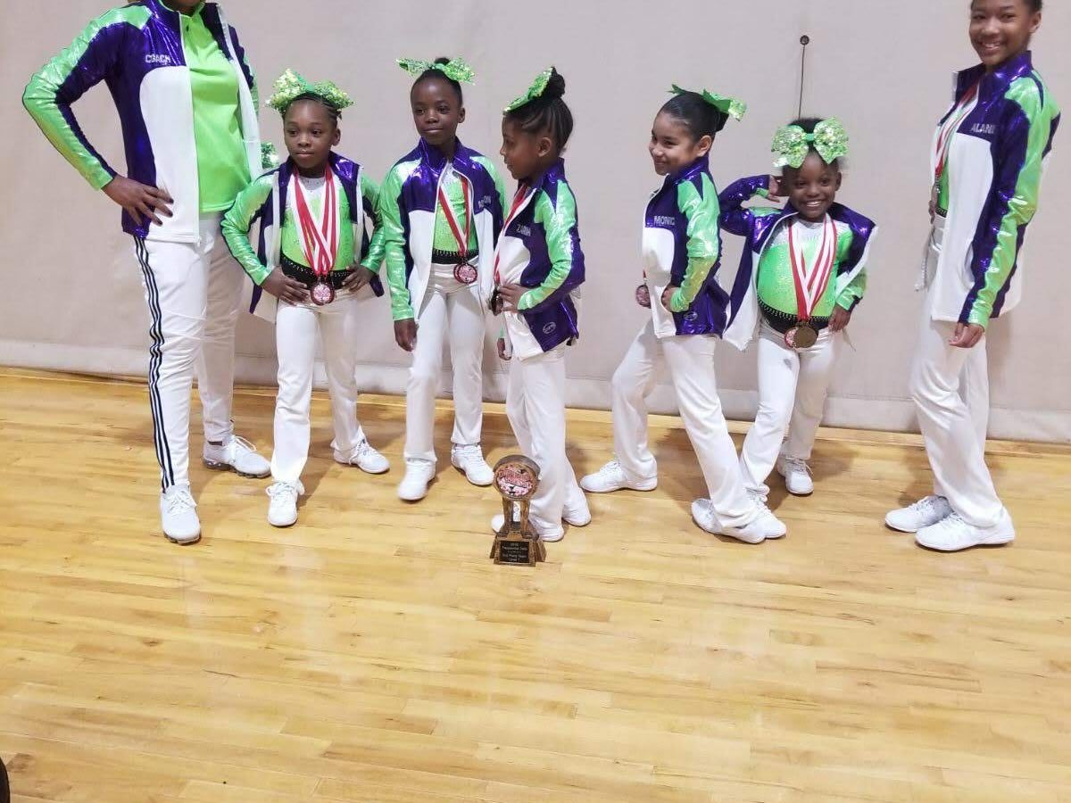 Six dancers in purple, green, and white costumes posing in a studio with a wooden floor