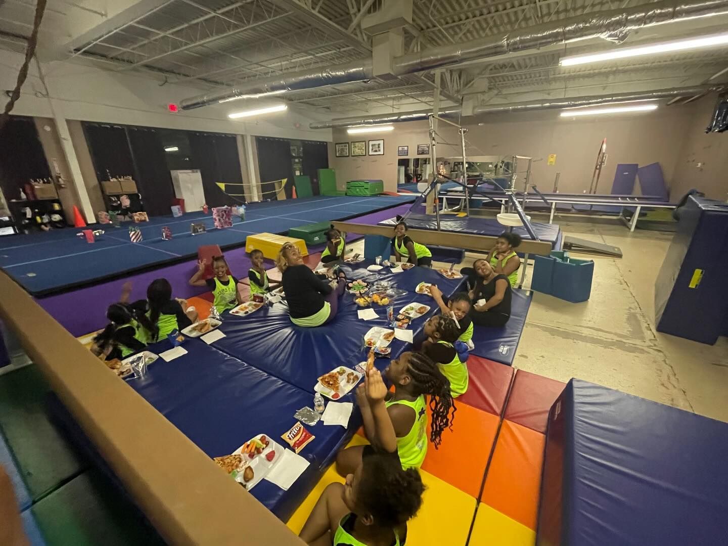 Children seated on blue mats in a gym, eating snacks at a long table during a group activity.
