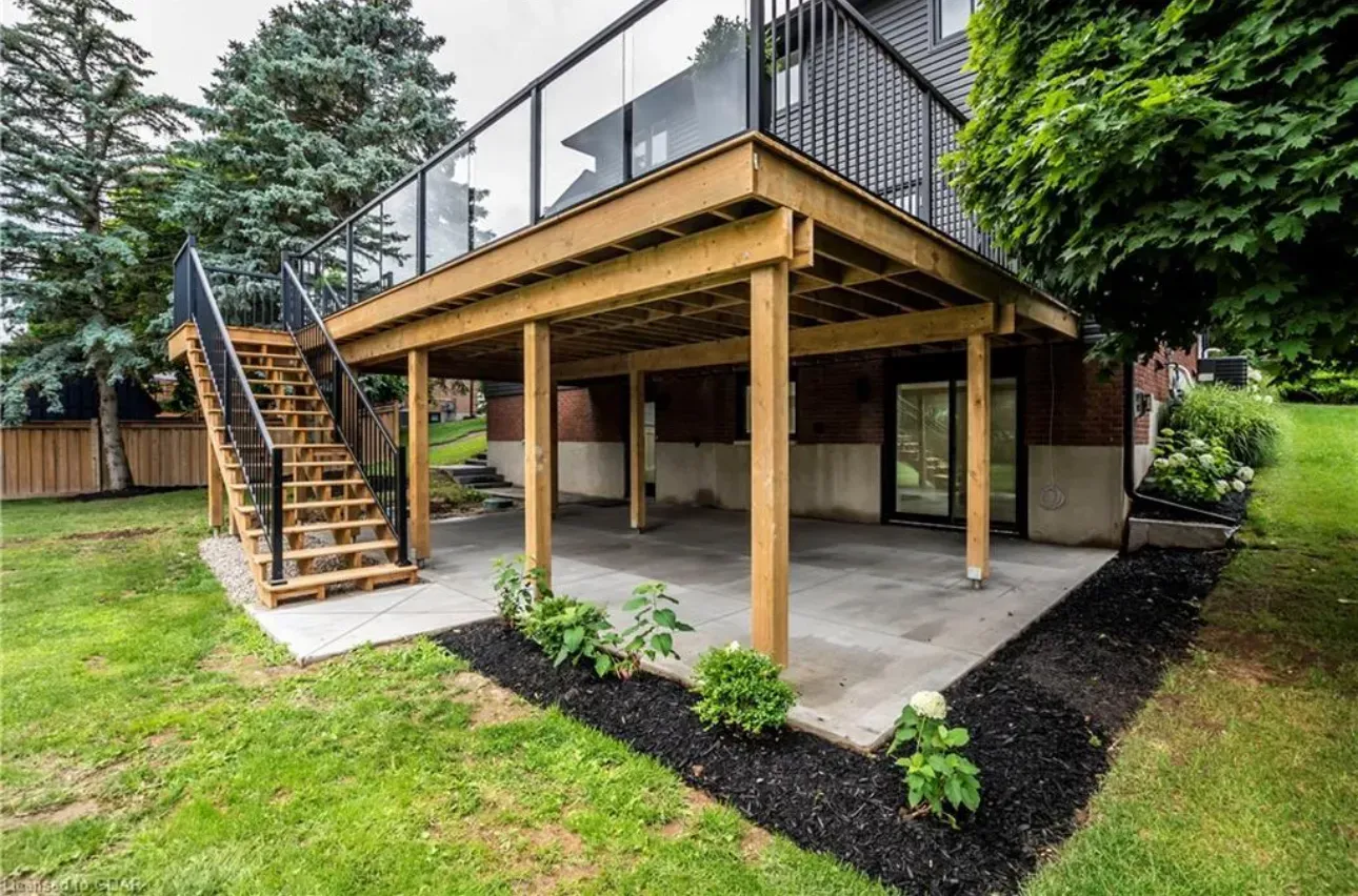 Wooden deck with black railing and stairs over a concrete patio; surrounded by grass and landscaping.