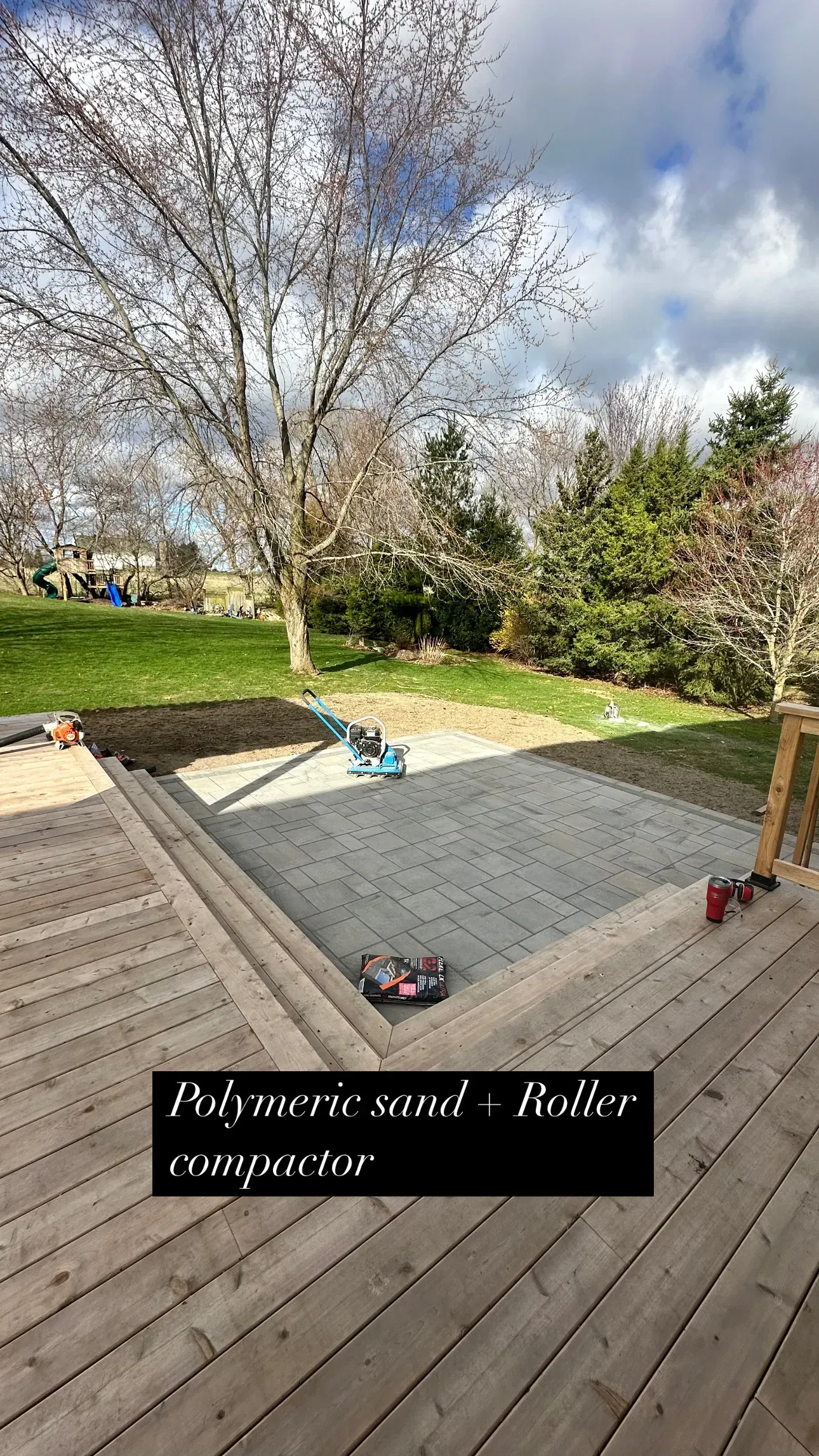 A patio under construction, enclosed by a wooden deck, with a gravel base. Green lawn and trees in the background.