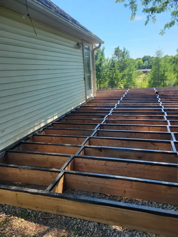 Deck framework construction next to a house with horizontal siding. Brown wood, black flashing. Outdoors, sunny.
